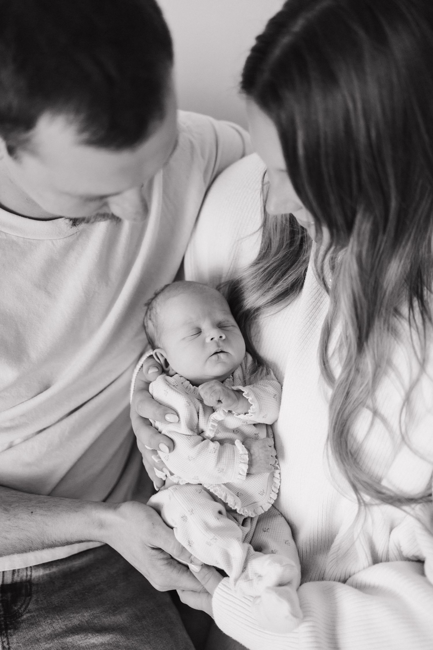 Black and white photo of new parents lovingly gazing at their sleeping newborn baby cradled in their arms.