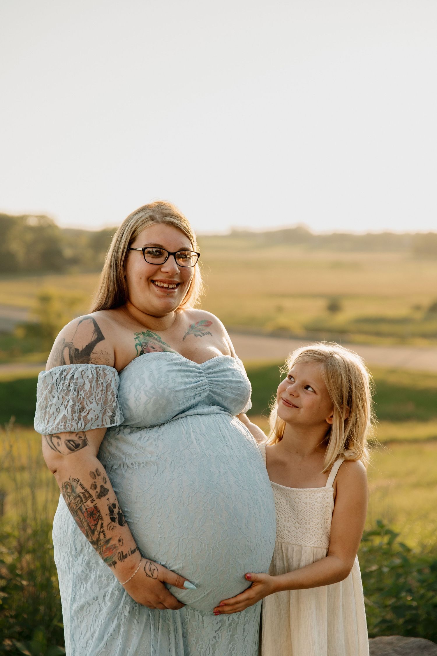Pregnant woman in blue lace dress with tattoos smiles outdoors as young blonde girl in white dress touches her baby bump.