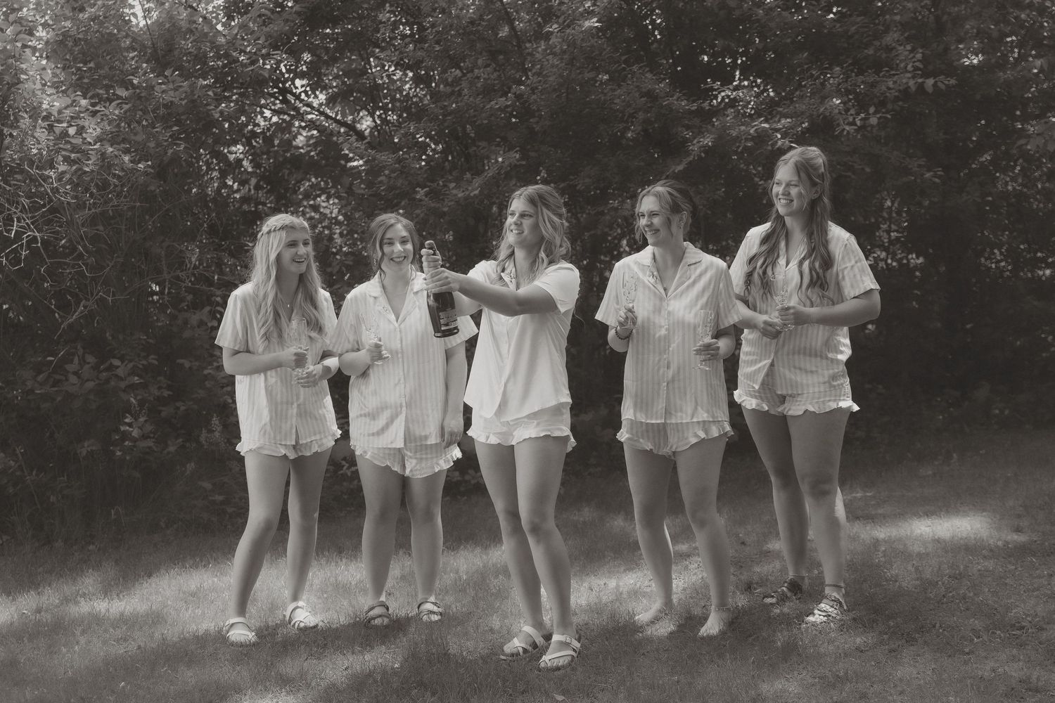 Five young women in matching white shorts and shirts laugh together outdoors in a black and white photo.