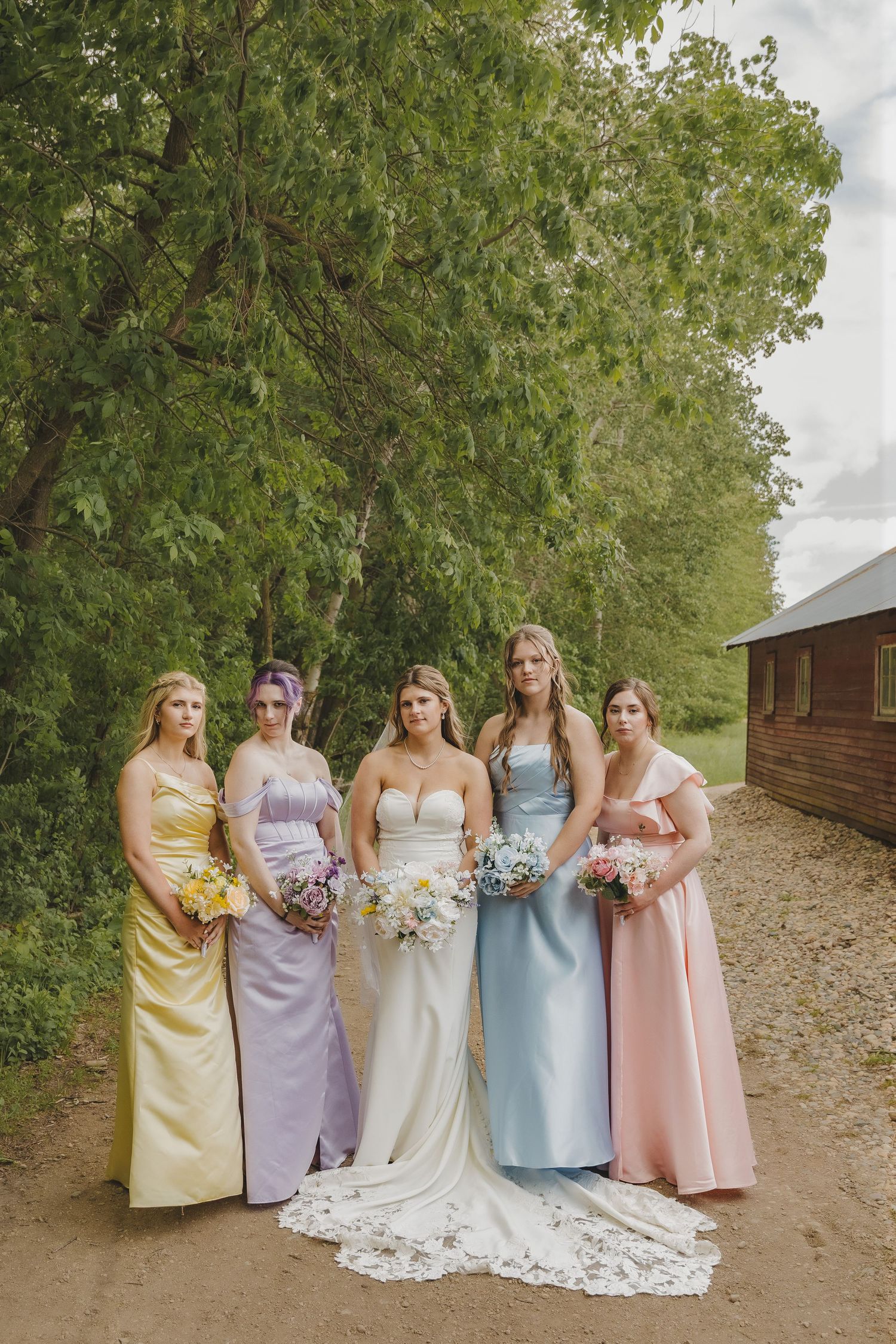 A bride in a white strapless gown stands with four bridesmaids in pastel dresses holding bouquets outdoors.