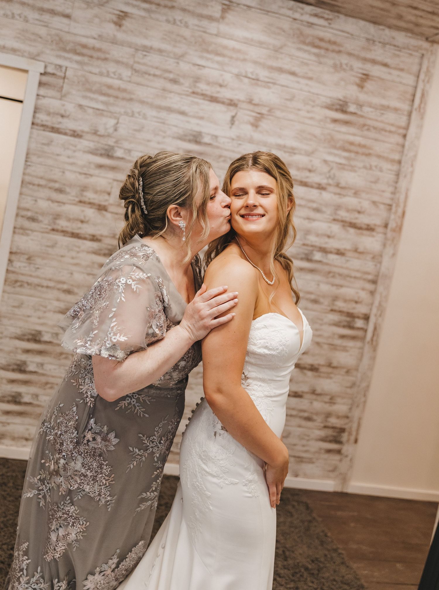 A smiling bride in a white strapless gown receives a kiss on the cheek from a woman in a silver lace dress.
