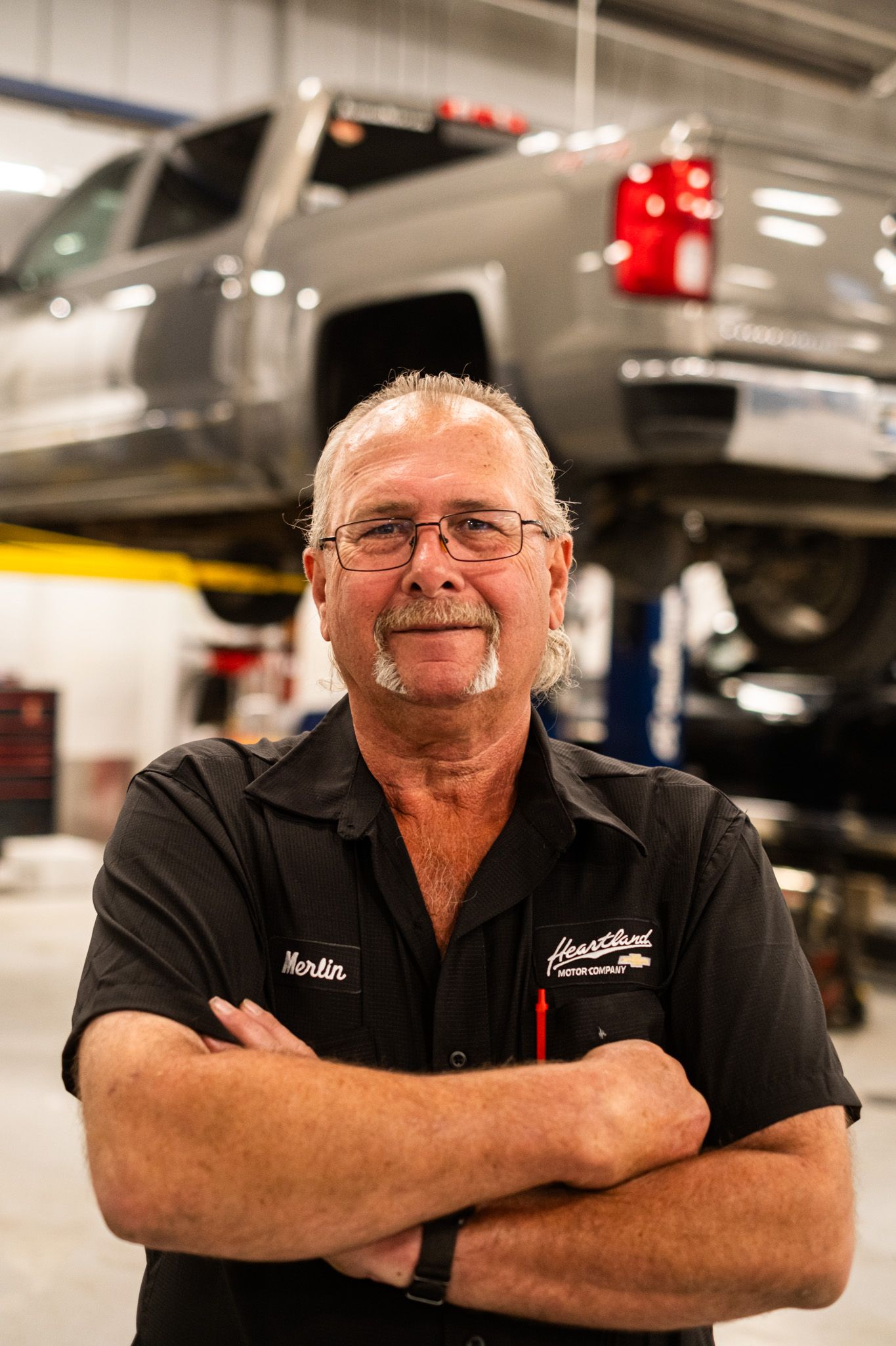 Auto service professional in black uniform standing confidently in repair shop with lifted truck in background.
