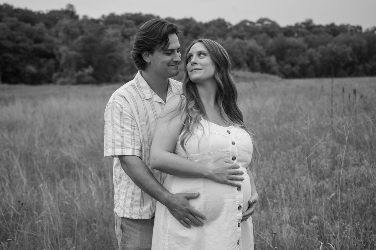 A black and white maternity photo captures a tender moment between expectant parents in a field.