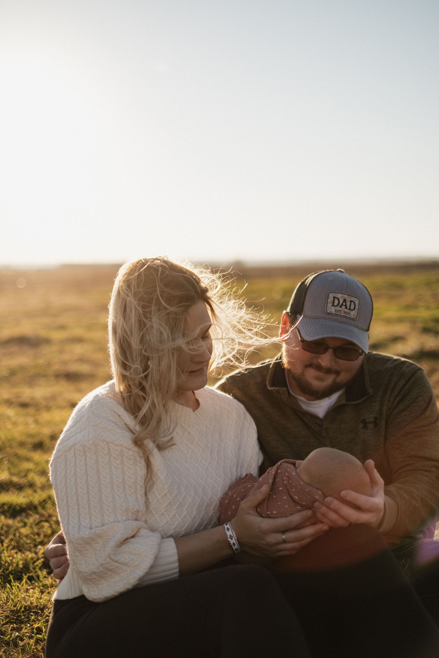Couple sitting in a sunlit field at golden hour holding their newborn baby.
