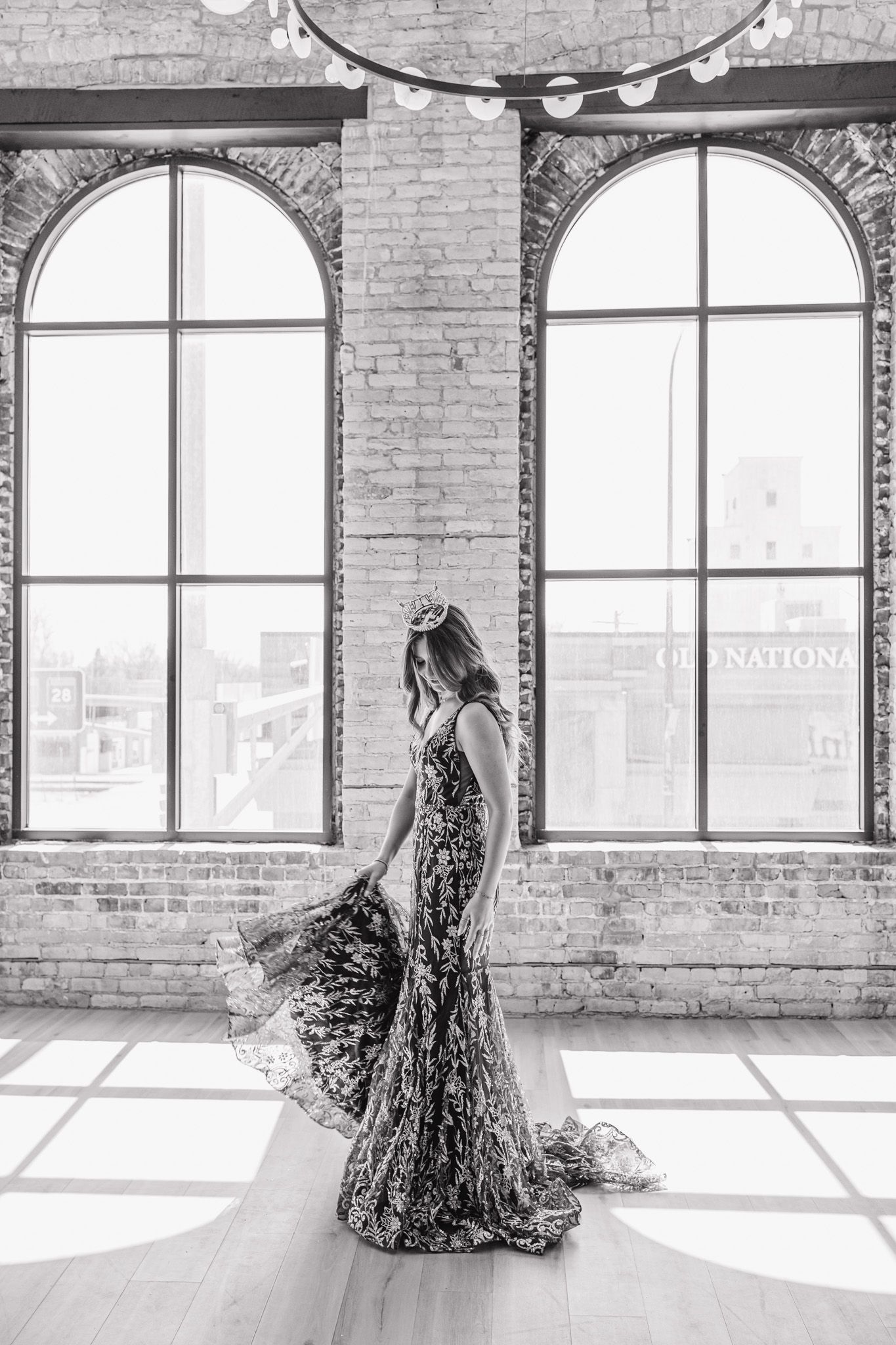 Black and white photo of a woman in a sequined gown and crown twirling in a sunlit brick loft.