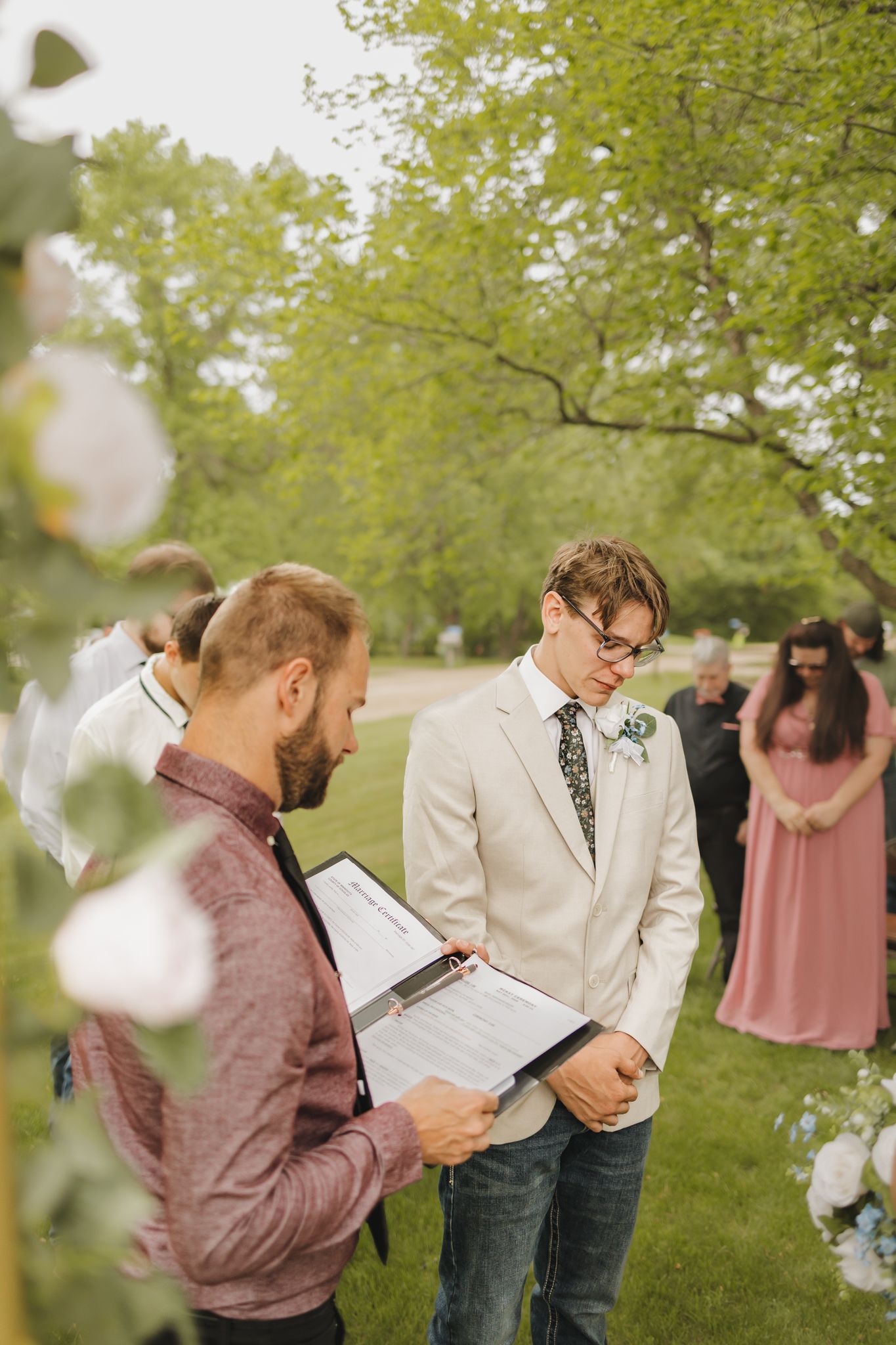 Groom in beige suit reads vows with officiant during outdoor wedding ceremony surrounded by guests and lush green trees.