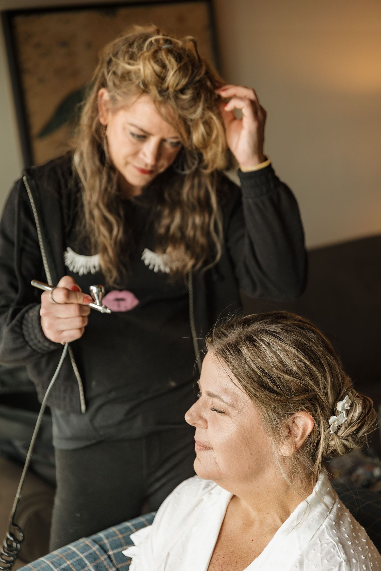A makeup artist applies airbrush foundation to a bride in a white robe, preparing her for her wedding day.
