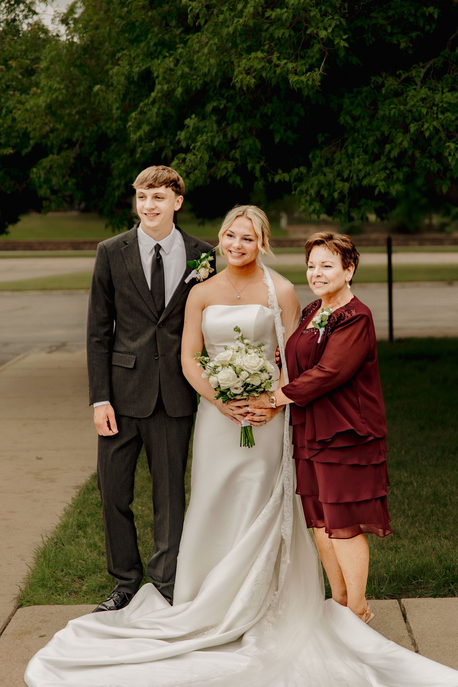 Blonde bride in white strapless gown holding white bouquet posing with young man and older woman outdoors.