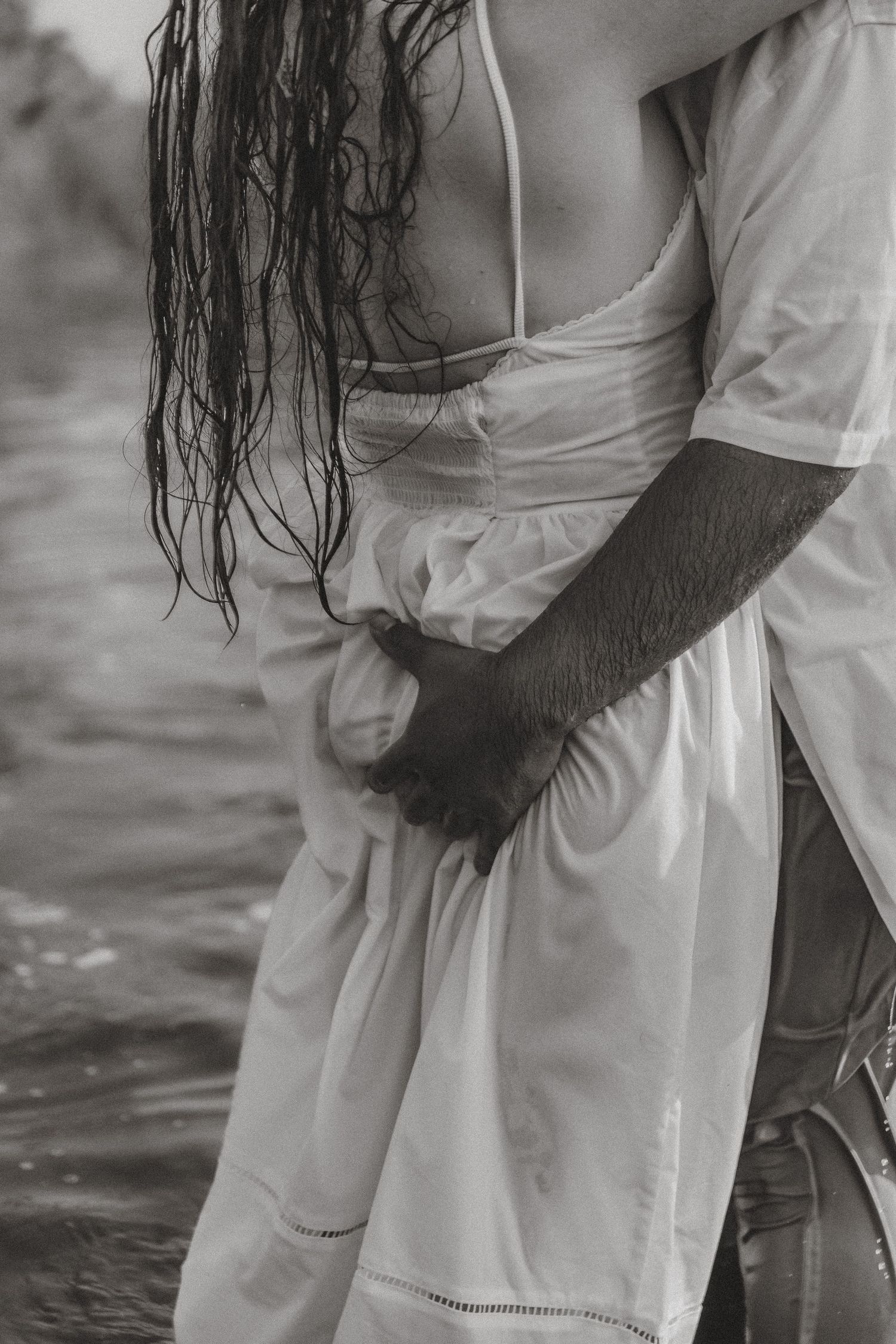 Black and white photo of a couple embracing near water, woman in white dress with wet hair, man's arm wrapped around her waist.