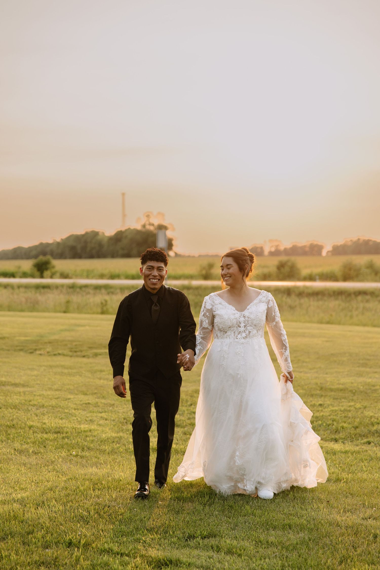 A couple walks hand-in-hand at sunset, bride in lace gown and groom in black suit, on a green field.