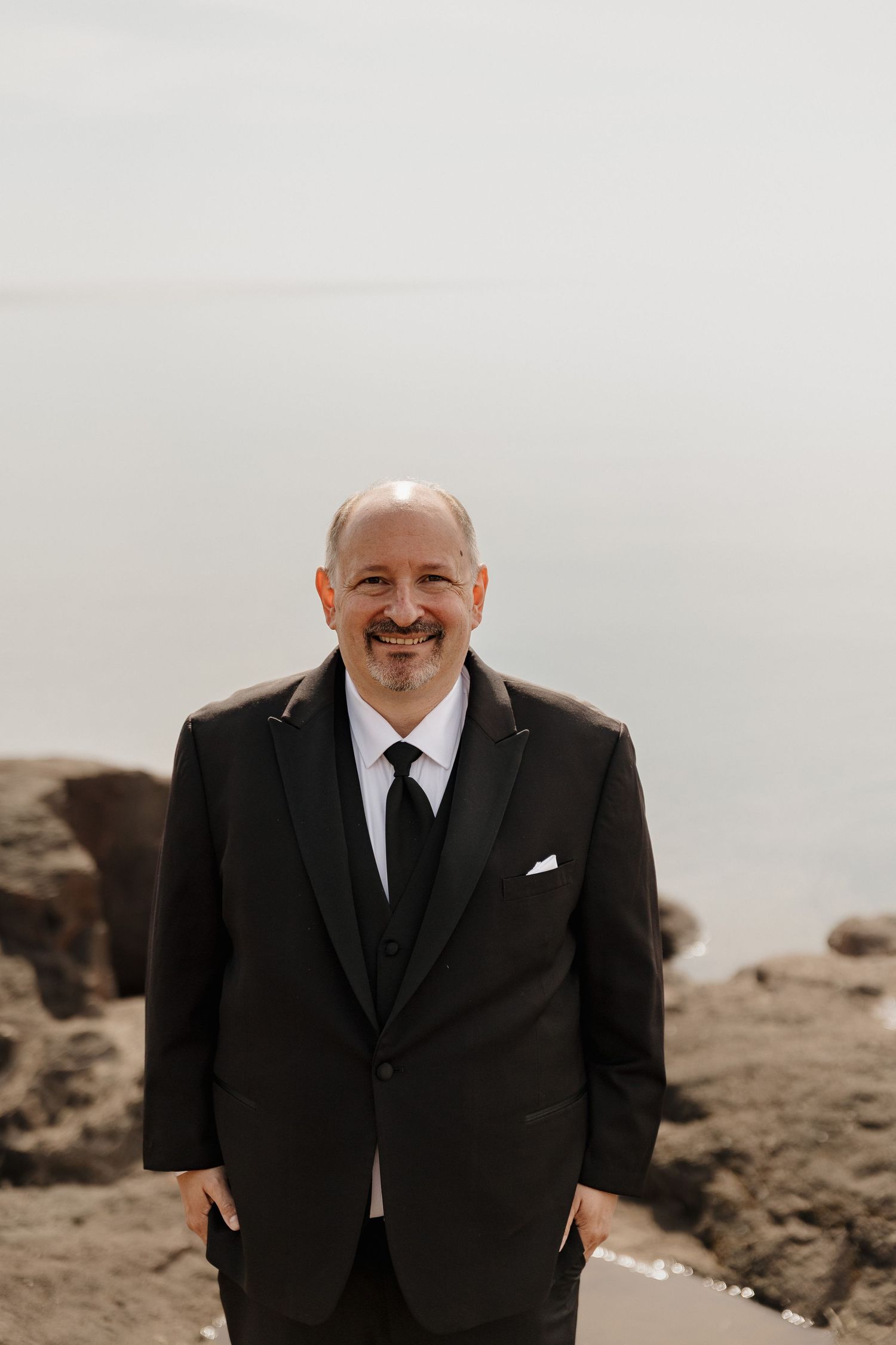 Smiling groom in black tuxedo standing on rocky shore with misty water behind him.
