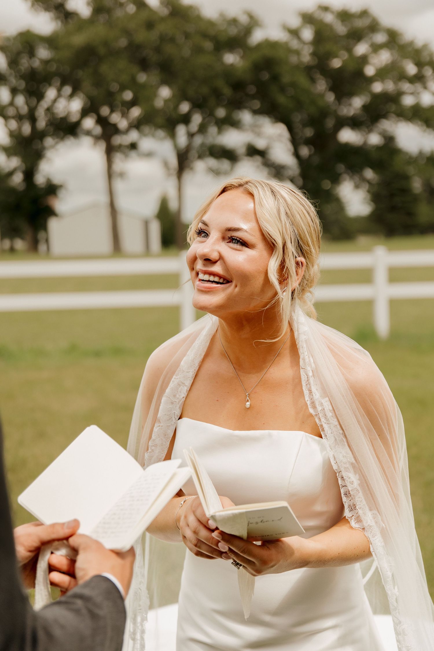 Smiling blonde bride in white strapless gown and veil holding books during outdoor wedding ceremony on green lawn.