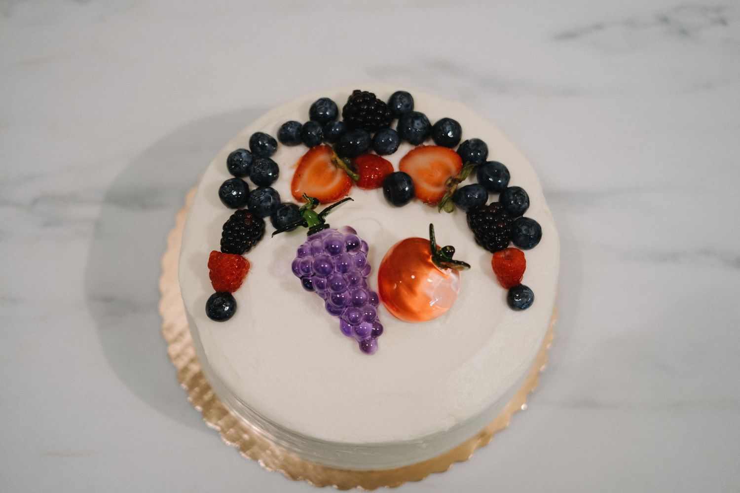 White frosted cake decorated with fresh berries and grapes on a marble surface.