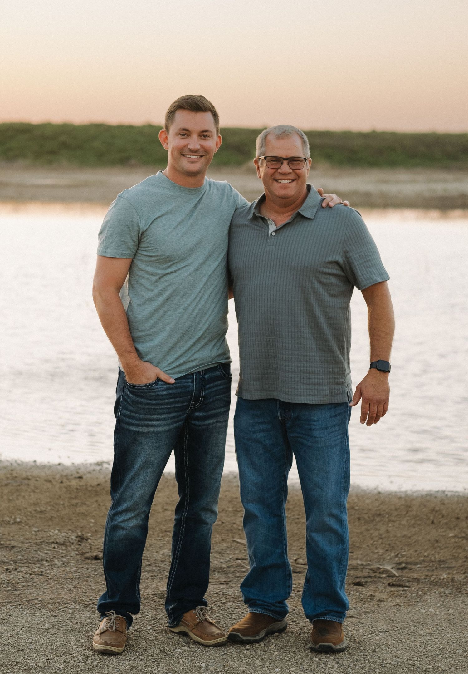 Two people in matching teal shirts and jeans pose together at sunset by the water.