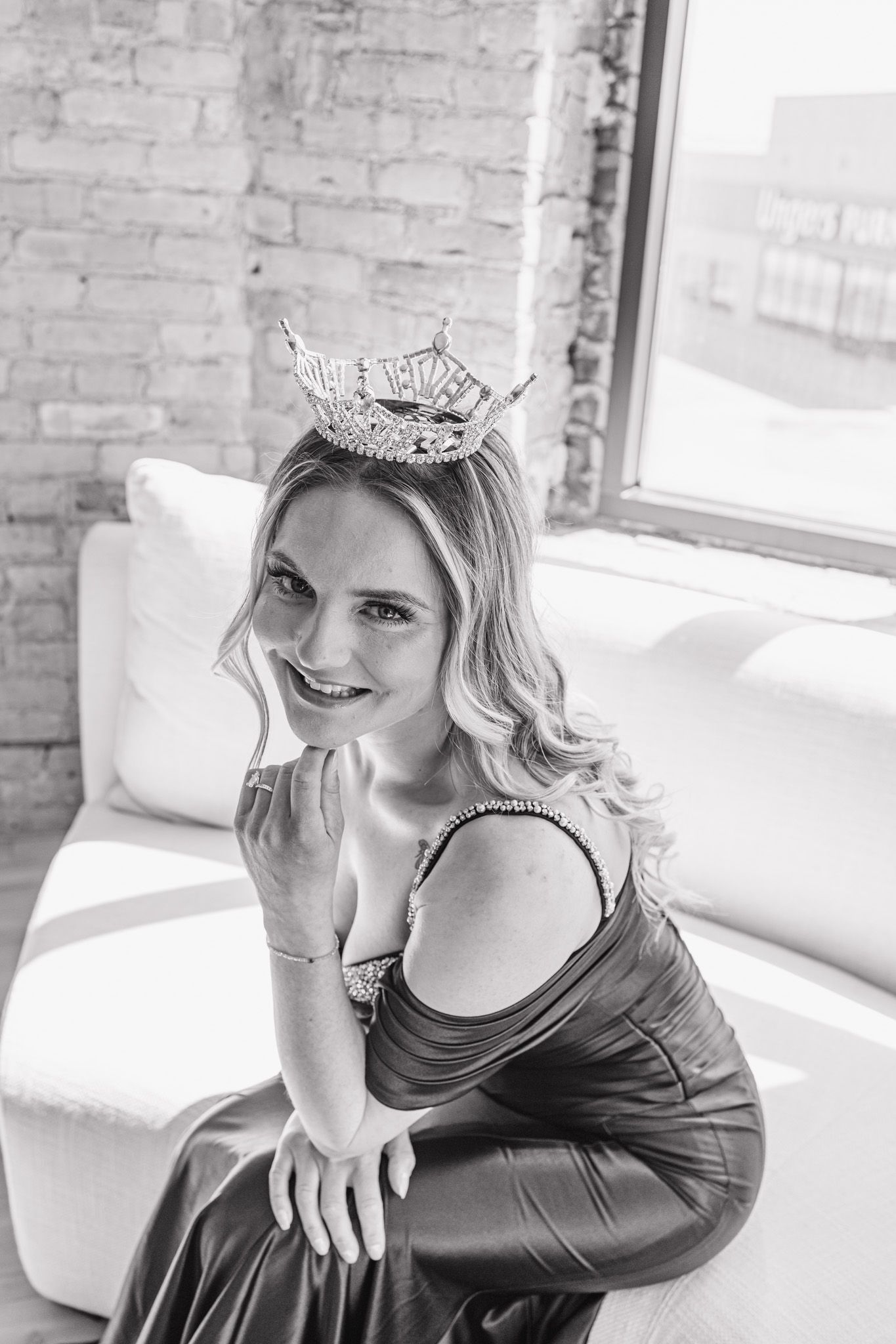 Smiling woman wearing a sparkling crown and dark gown seated on white sofa in brick-walled loft.