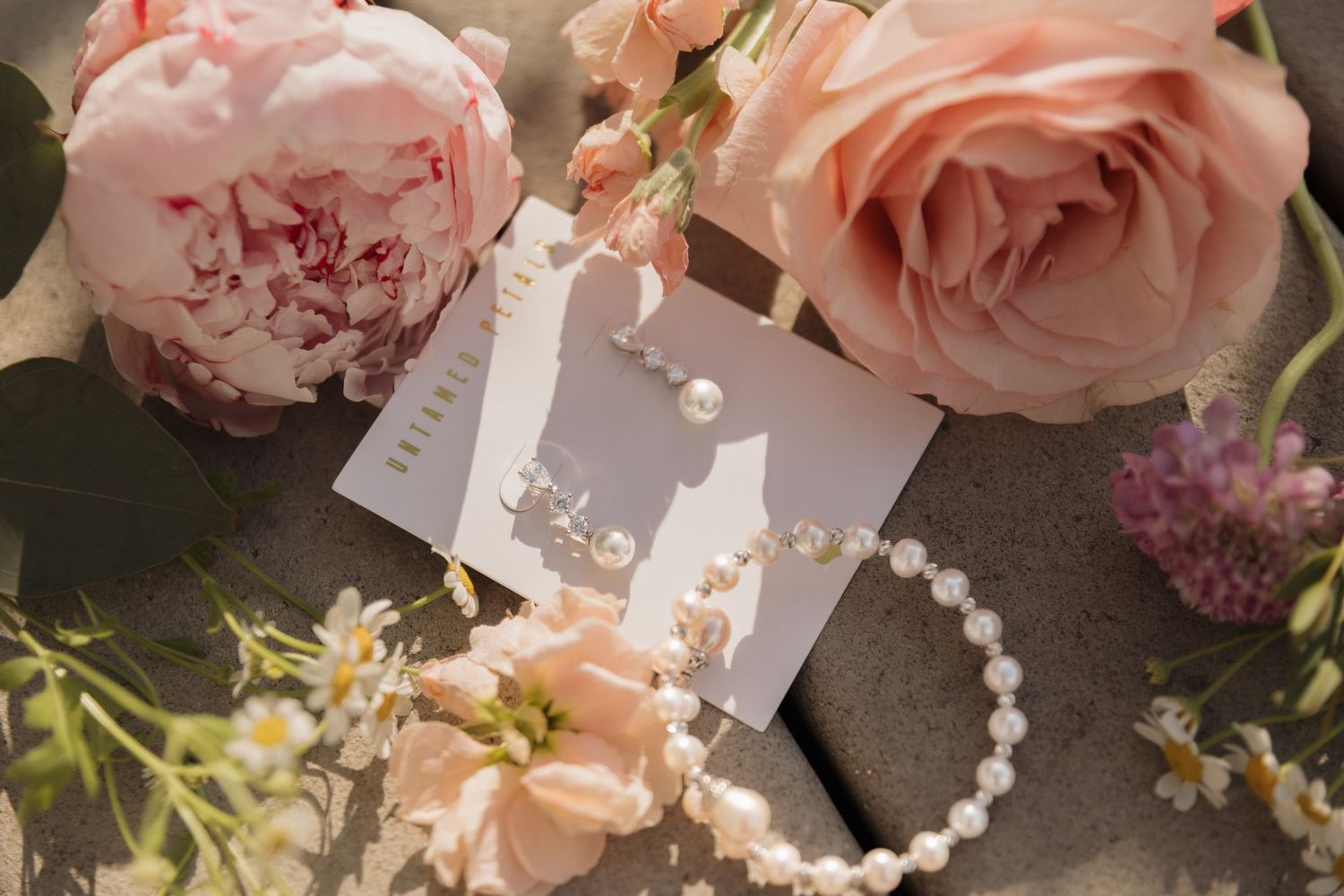 Pearl earrings and bracelet jewelry set displayed on card surrounded by soft pink roses and delicate wildflowers.