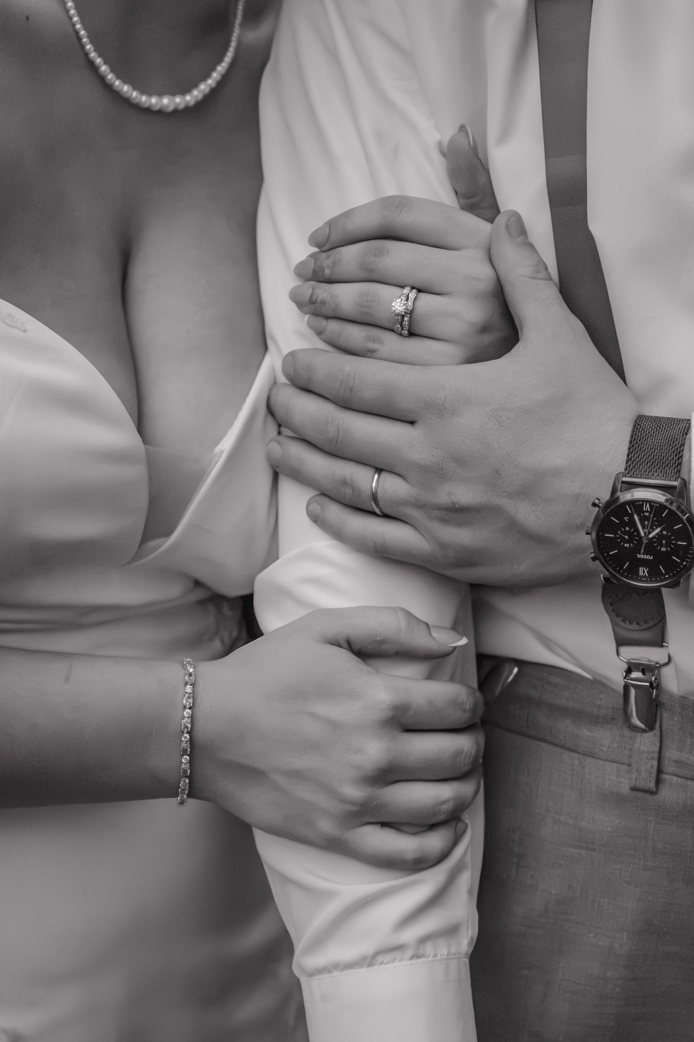 Black and white photo of newlyweds embracing, showing wedding rings, bracelet, and watch on intertwined hands.