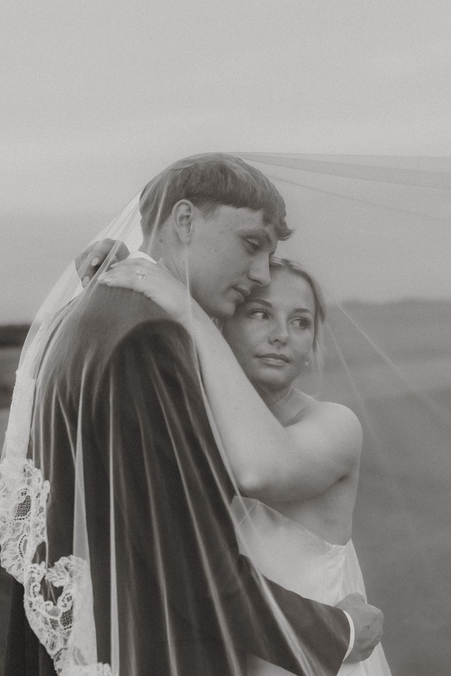 Black and white wedding photo of bride and groom embracing under a lace veil outdoors.