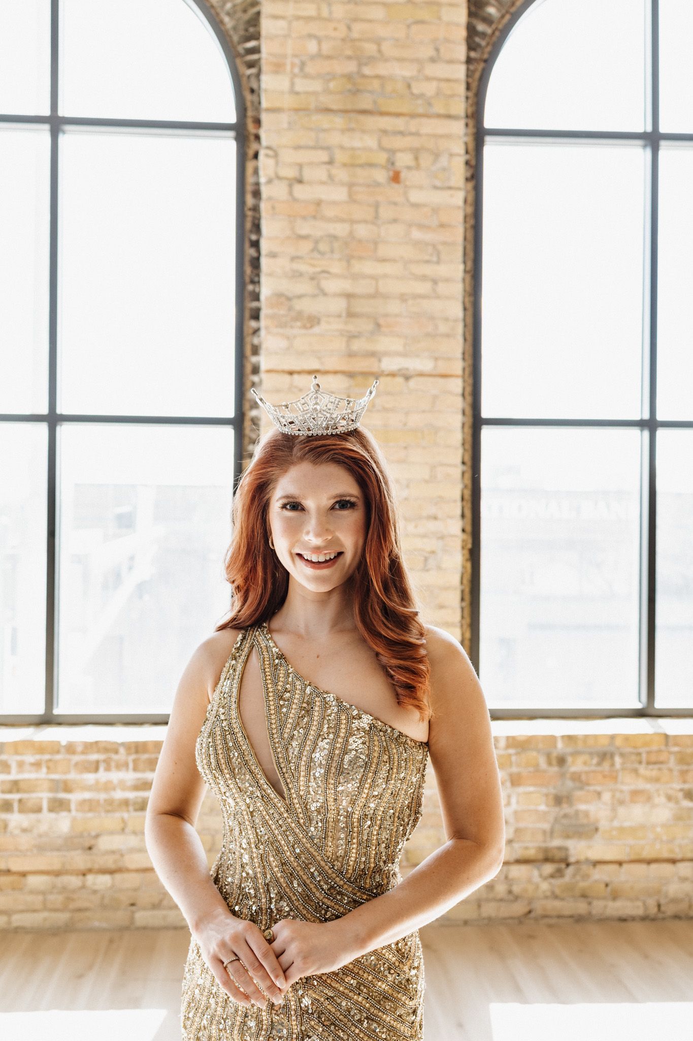 Smiling redhead woman wearing a gold sequined gown and diamond crown in a bright brick loft.