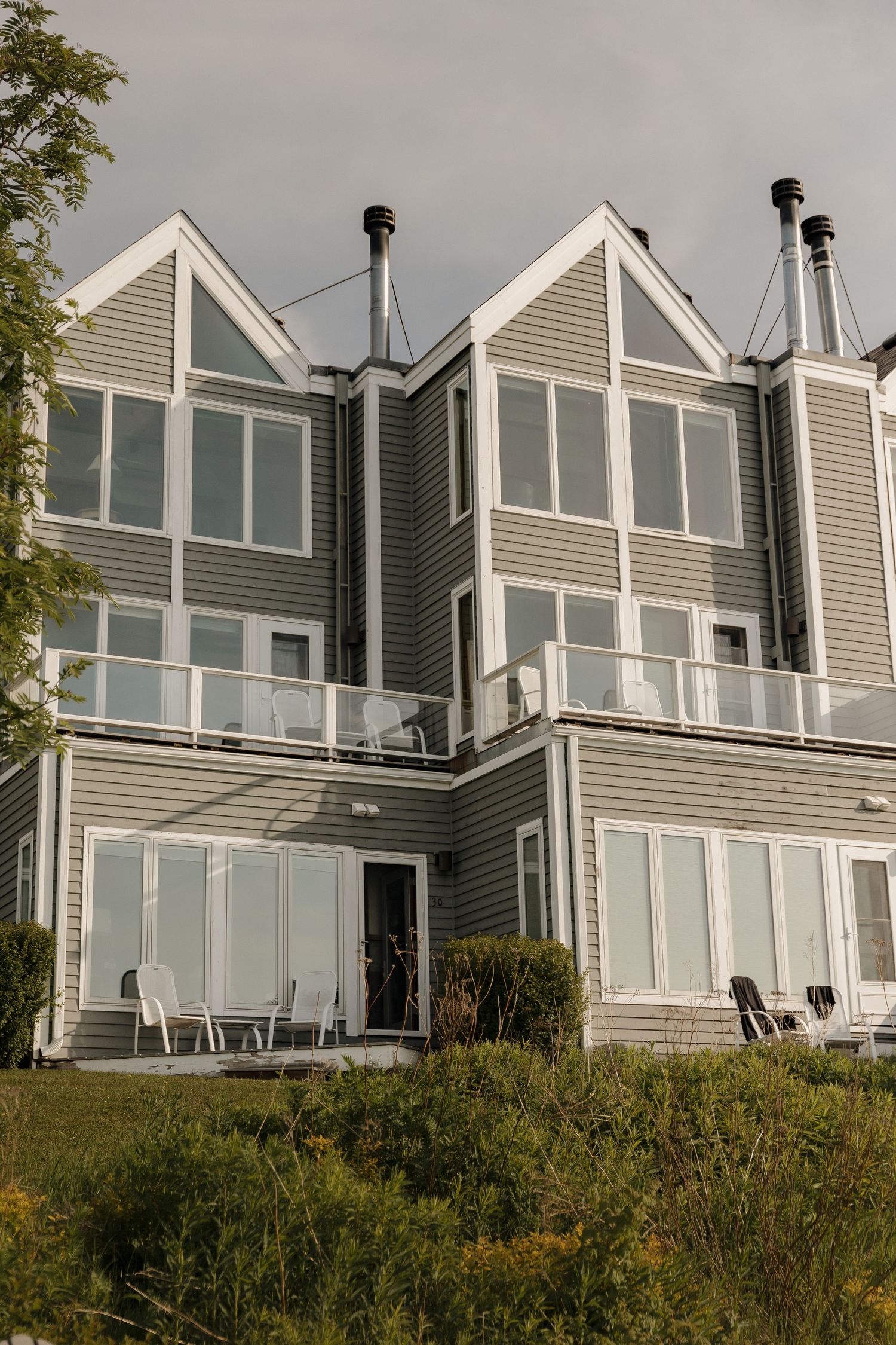 Gray multi-story coastal vacation home with large windows, white railings, and lush green landscaping under cloudy skies.