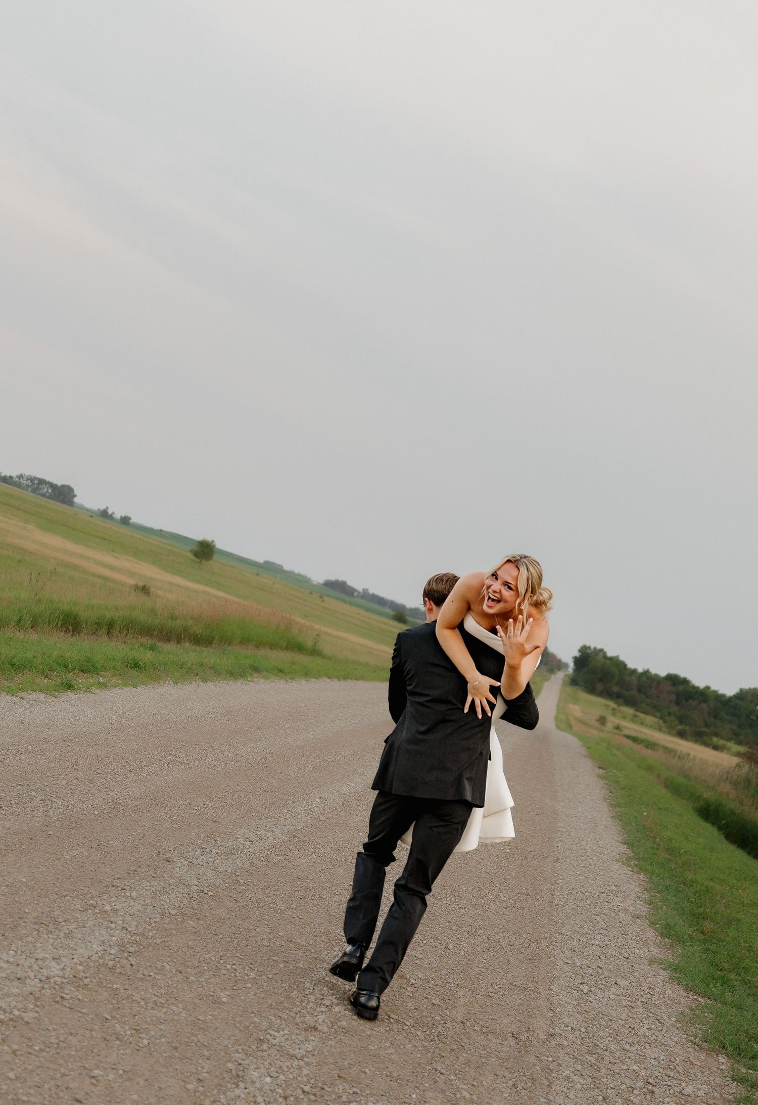 Groom lifting laughing bride on rural gravel road with open fields in background.