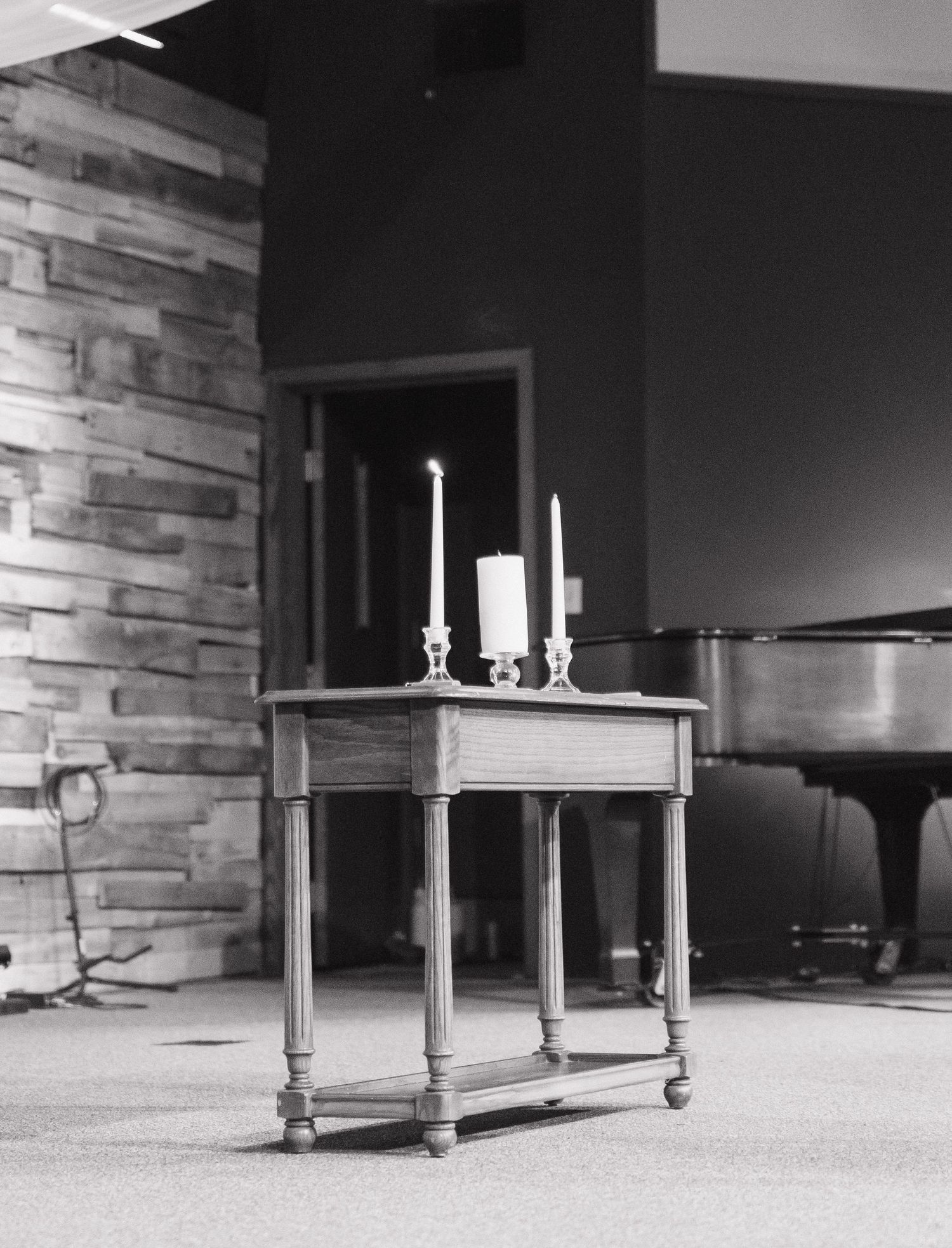 Black and white photo of small wooden table with candles near stone wall and grand piano at ceremony.