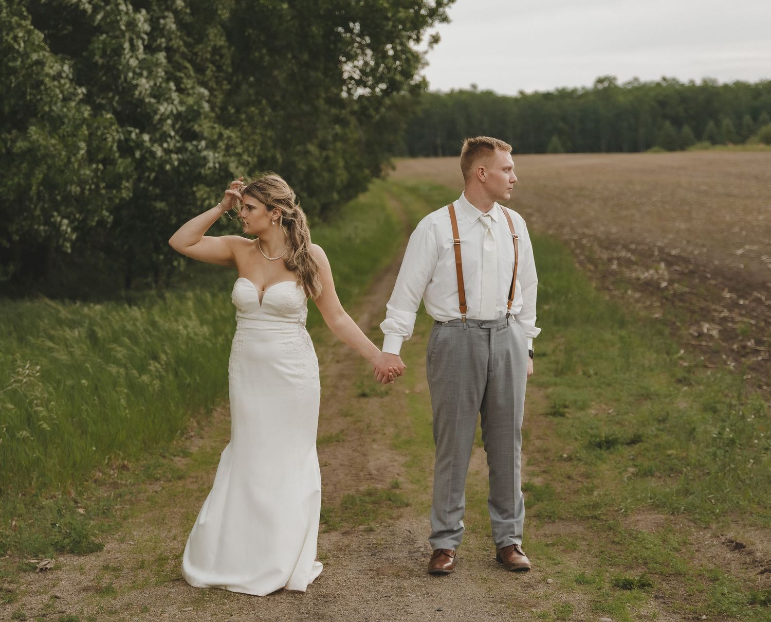 Bride and groom holding hands on a rustic dirt path surrounded by lush green fields on their wedding day.