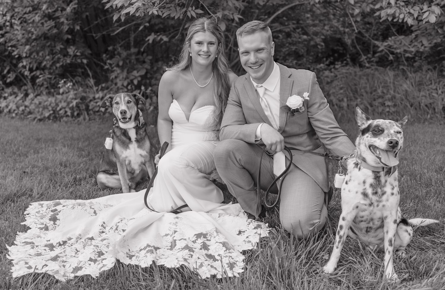 Black and white wedding photo of smiling couple sitting on grass with two spotted dogs outdoors.