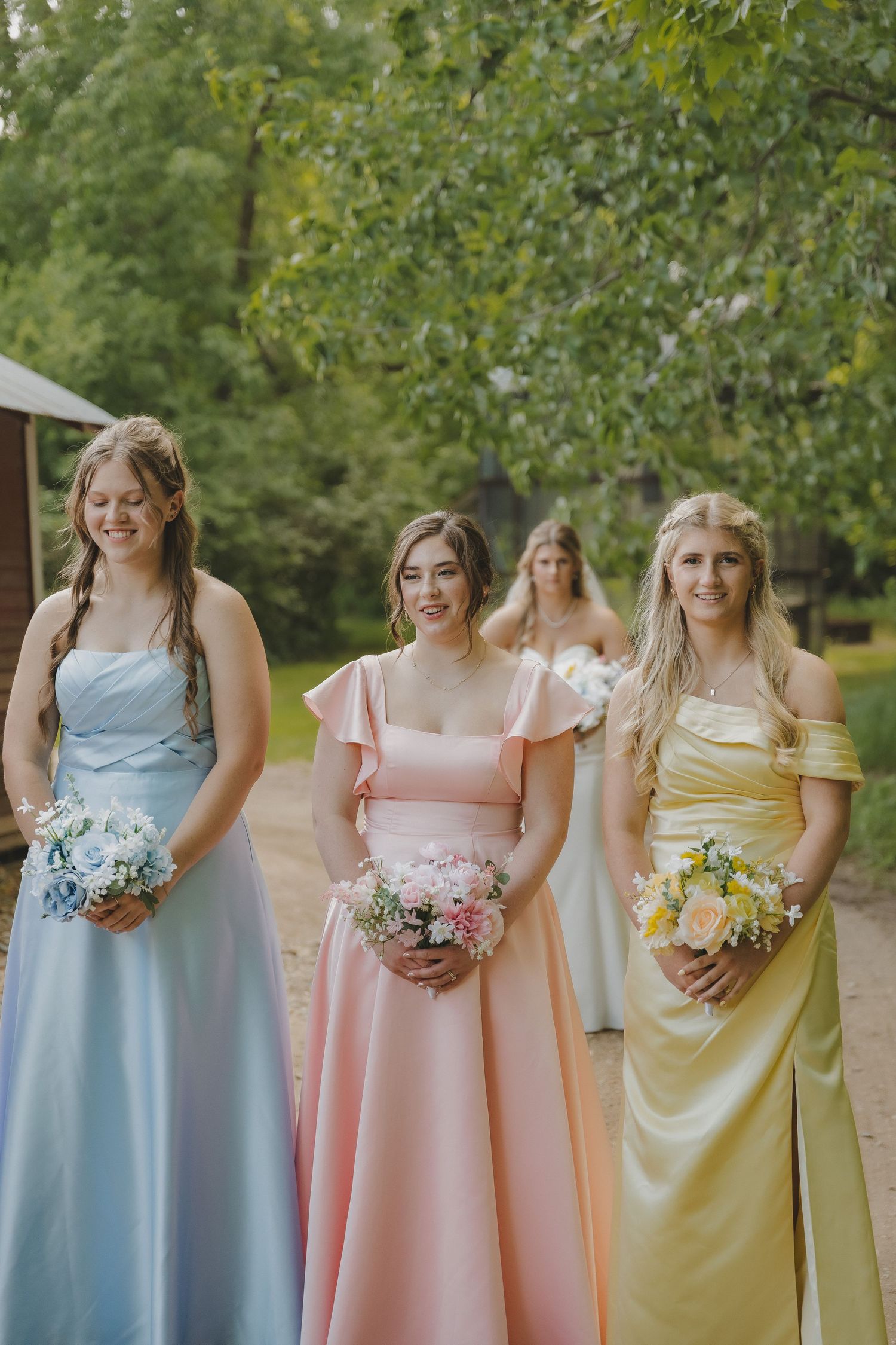 Three bridesmaids in pastel blue, pink, and yellow gowns holding bouquets outdoors.