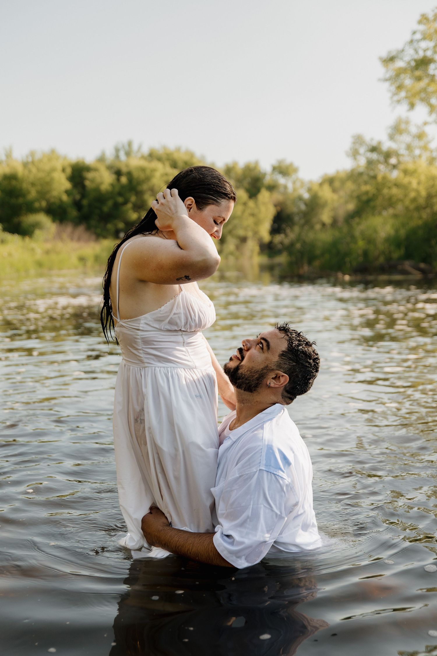A couple stands in a river, man kneeling in water gazing up at woman in white dress, surrounded by lush greenery.