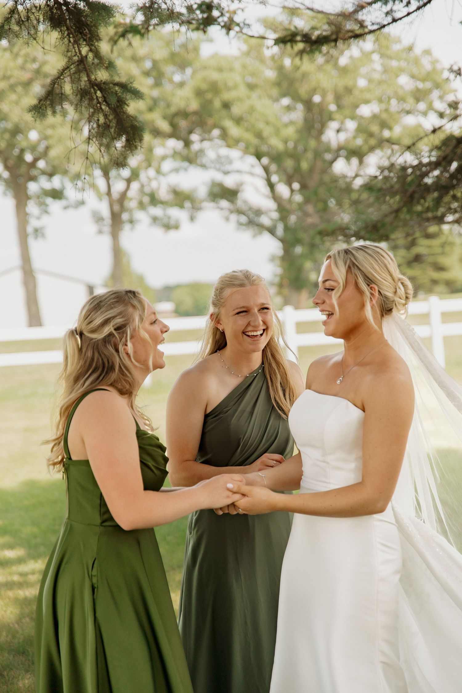 Bride in white strapless gown laughs joyfully with two bridesmaids in green dresses at outdoor wedding.