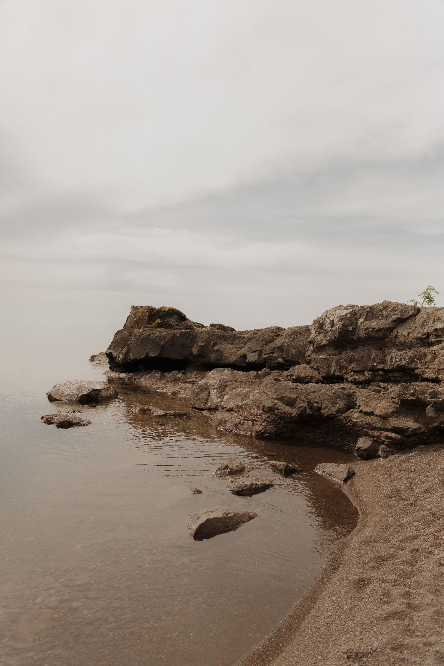 Rocky shoreline with calm water and sandy beach under overcast skies.