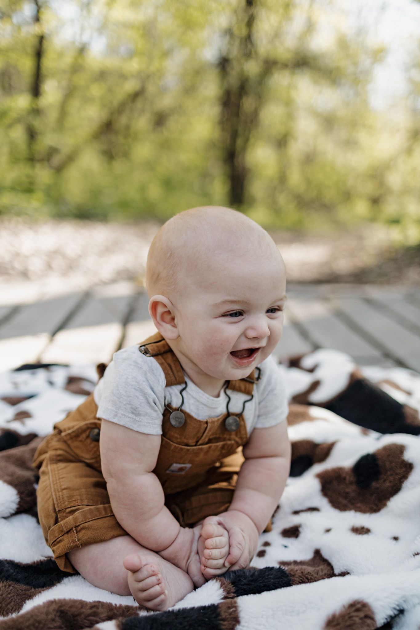 A happy baby in brown overalls sits on a cow-print blanket outdoors, smiling joyfully amid a lush green background.