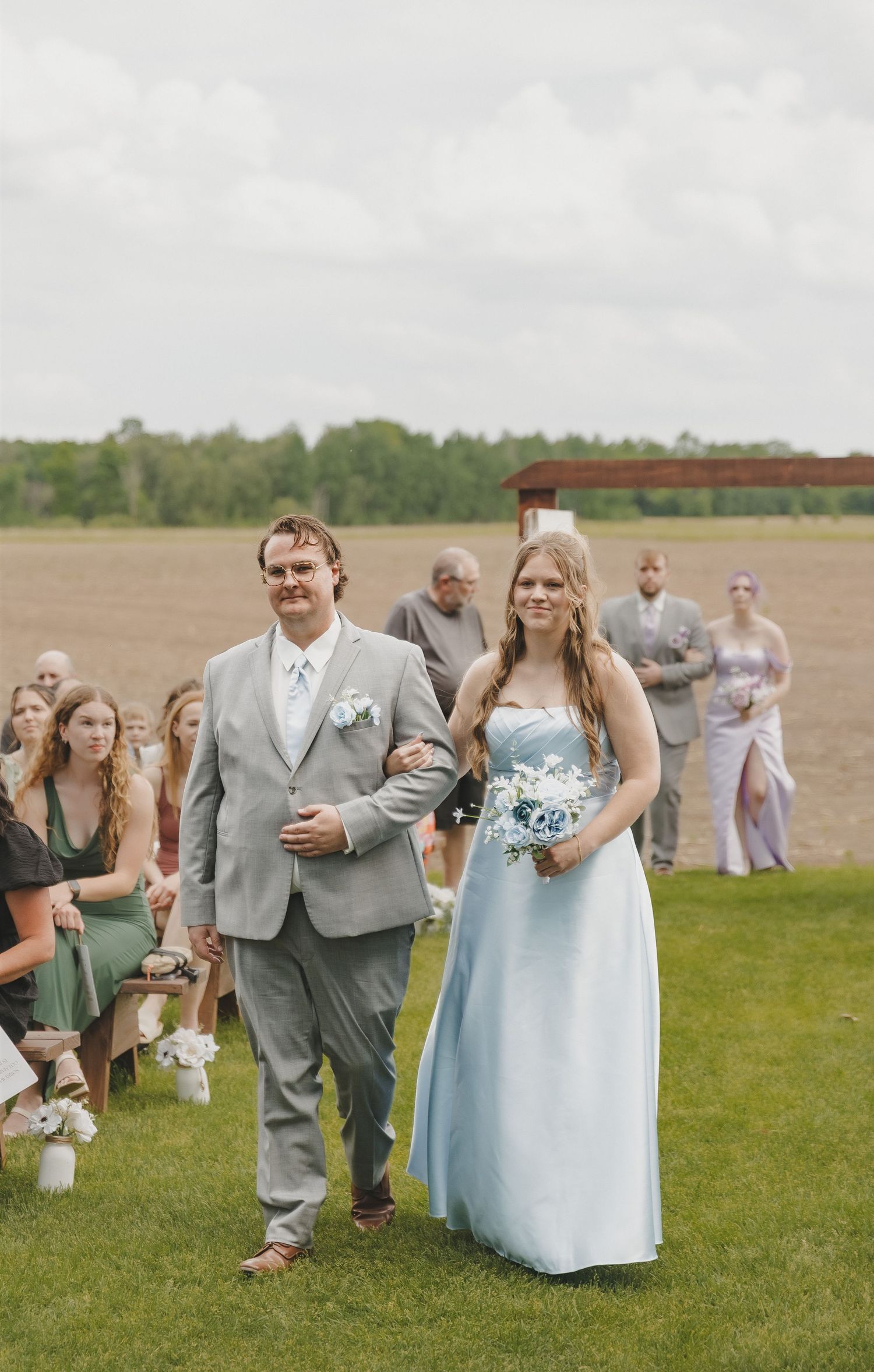 Groomsman escorts bridesmaid in light blue gown down outdoor wedding aisle on green lawn.