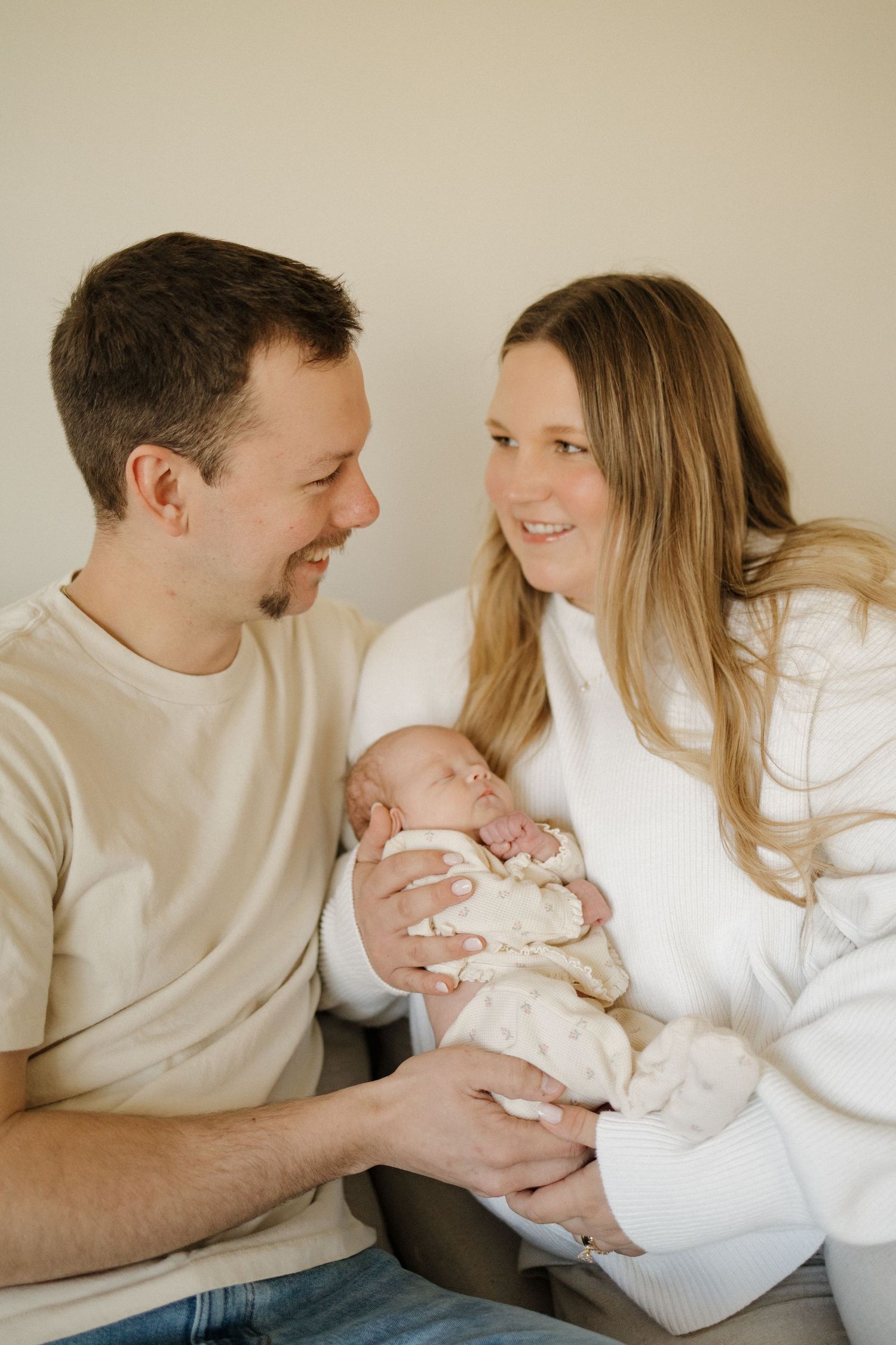 Smiling parents holding their newborn baby together, sharing a tender moment in a bright, neutral-toned room.