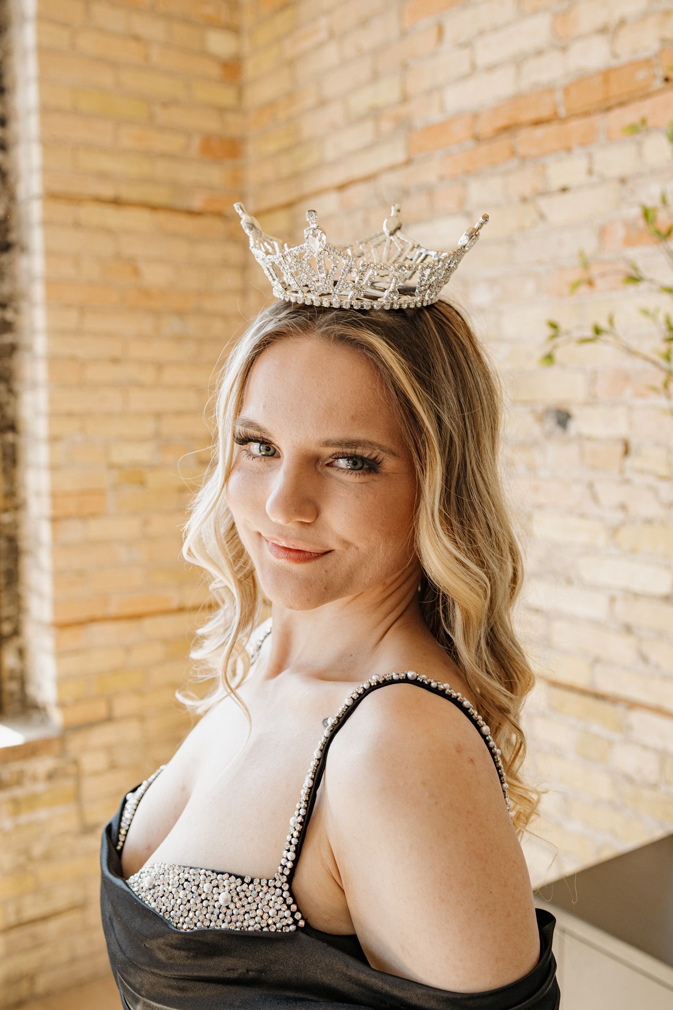 Blonde woman wearing a sparkling crown and black embellished gown poses elegantly in front of a brick wall.