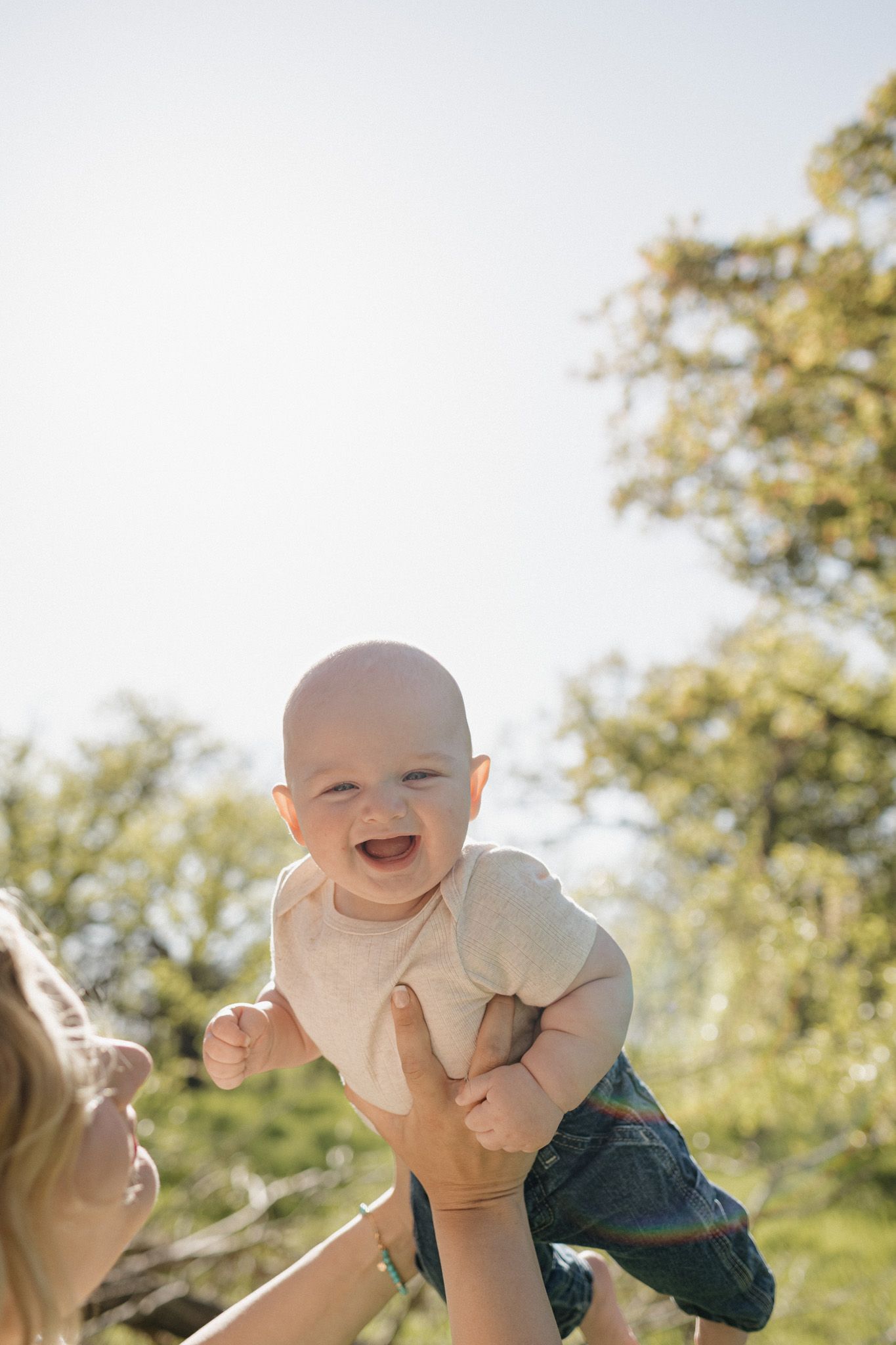 Joyful baby laughing while being lifted up outdoors in sunlight, with green trees in the background.