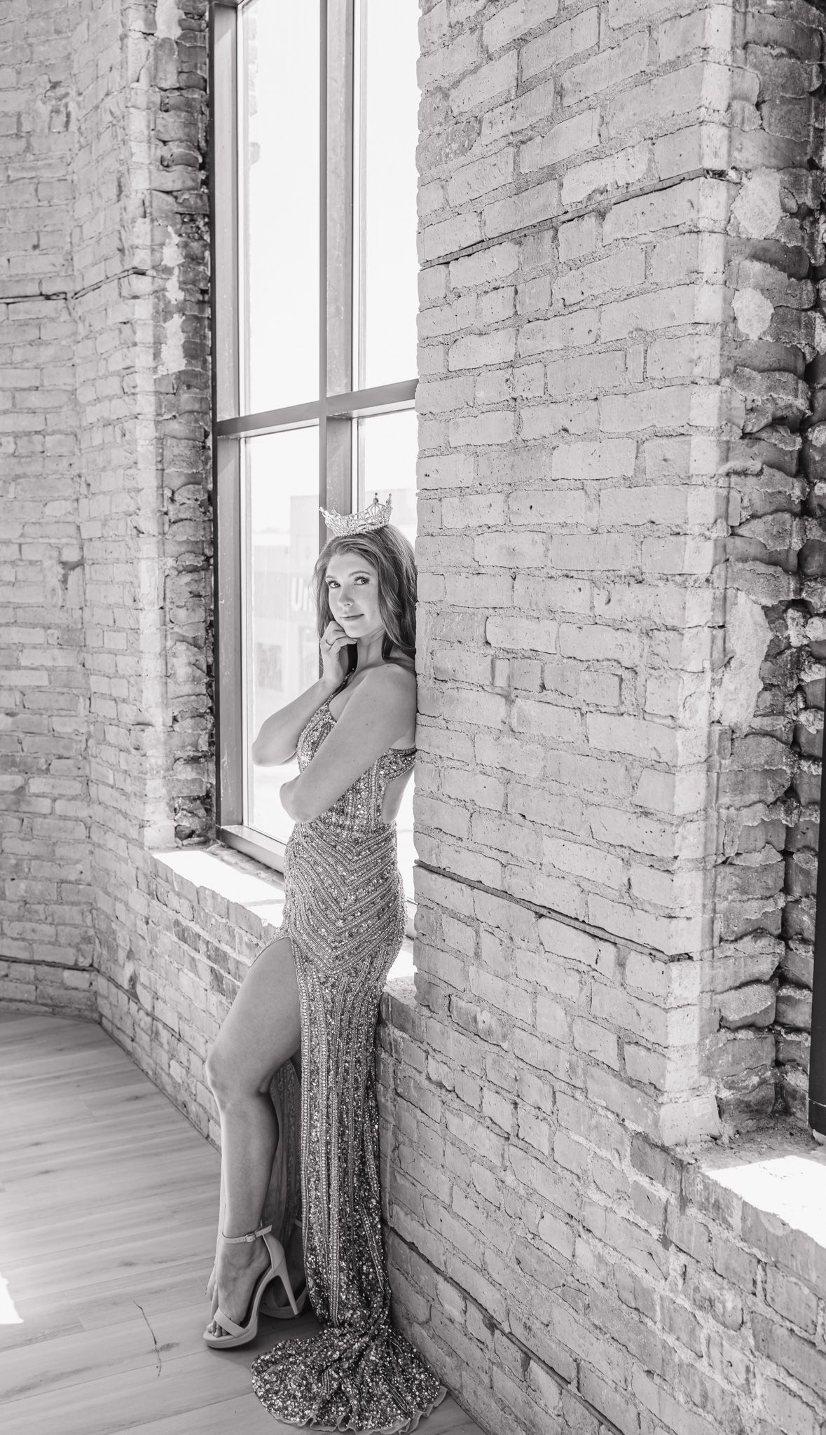 Woman in sequined gown poses by brick wall window in black and white portrait.