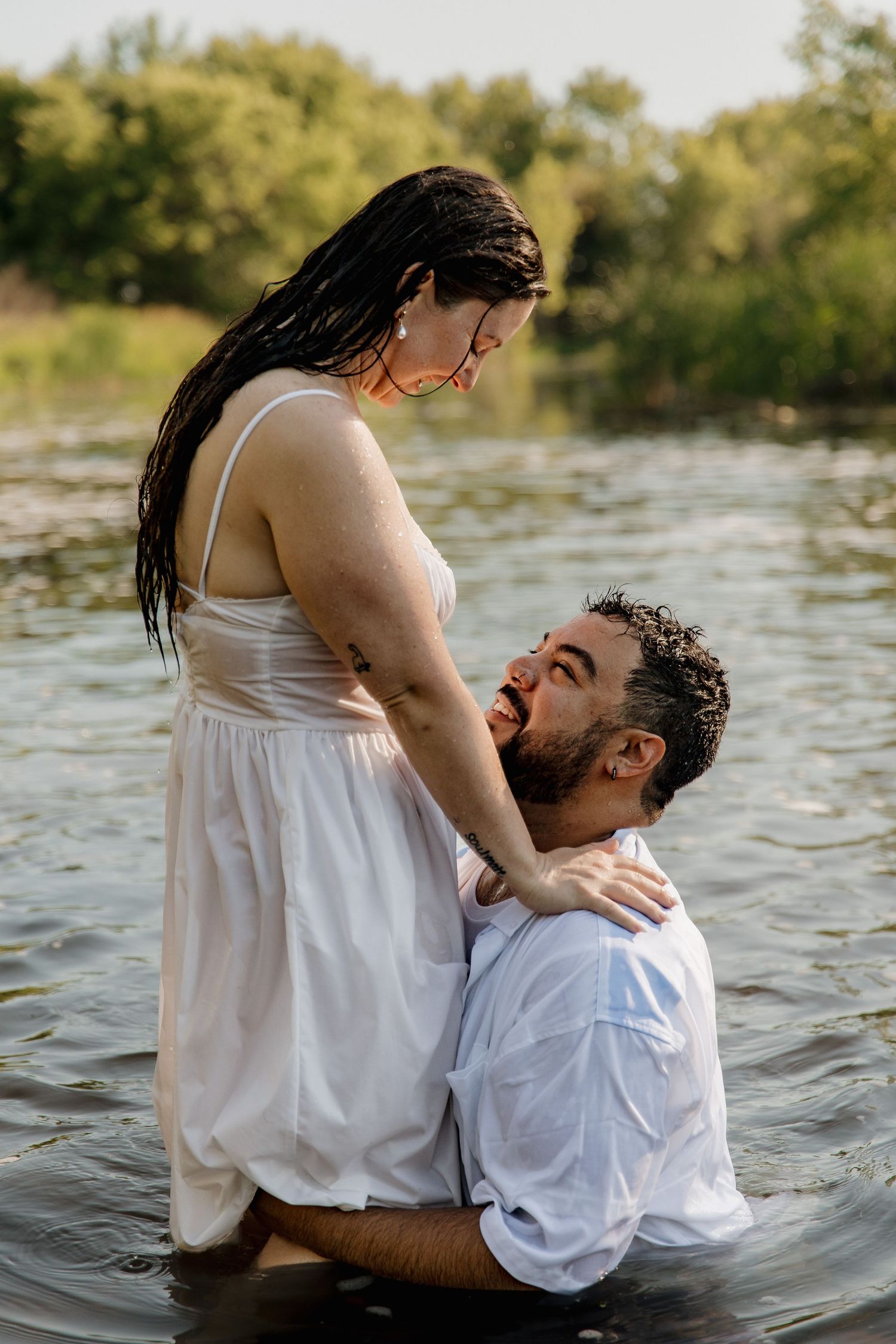 Couple in white outfits standing in a lake, sharing a romantic moment surrounded by lush green trees.
