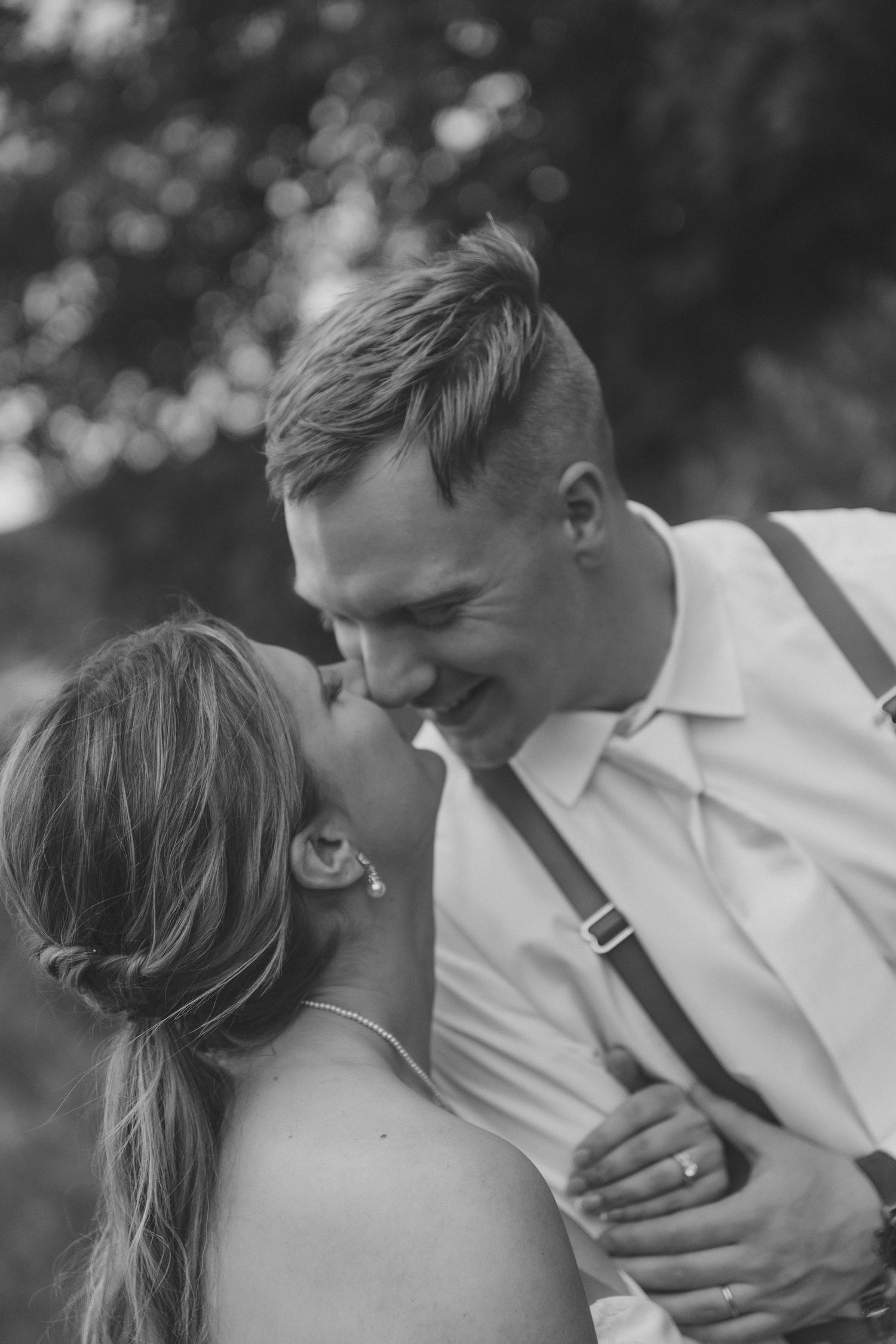 Black and white photo of a smiling groom in suspenders sharing a romantic kiss with his bride outdoors.