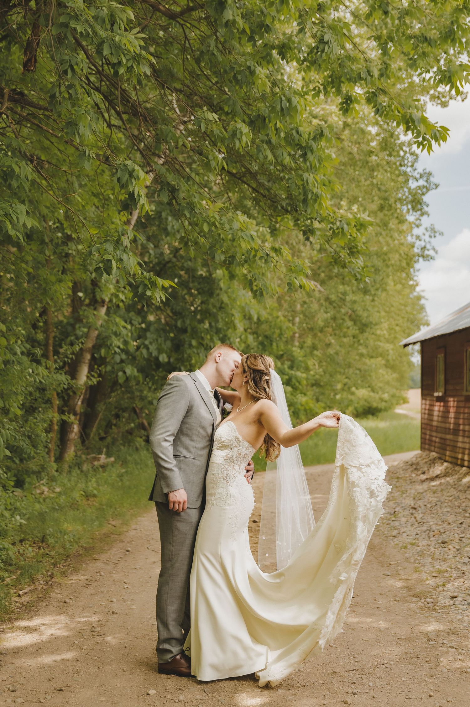 Bride and groom kissing on a dirt path with flowing veil and lush green trees.