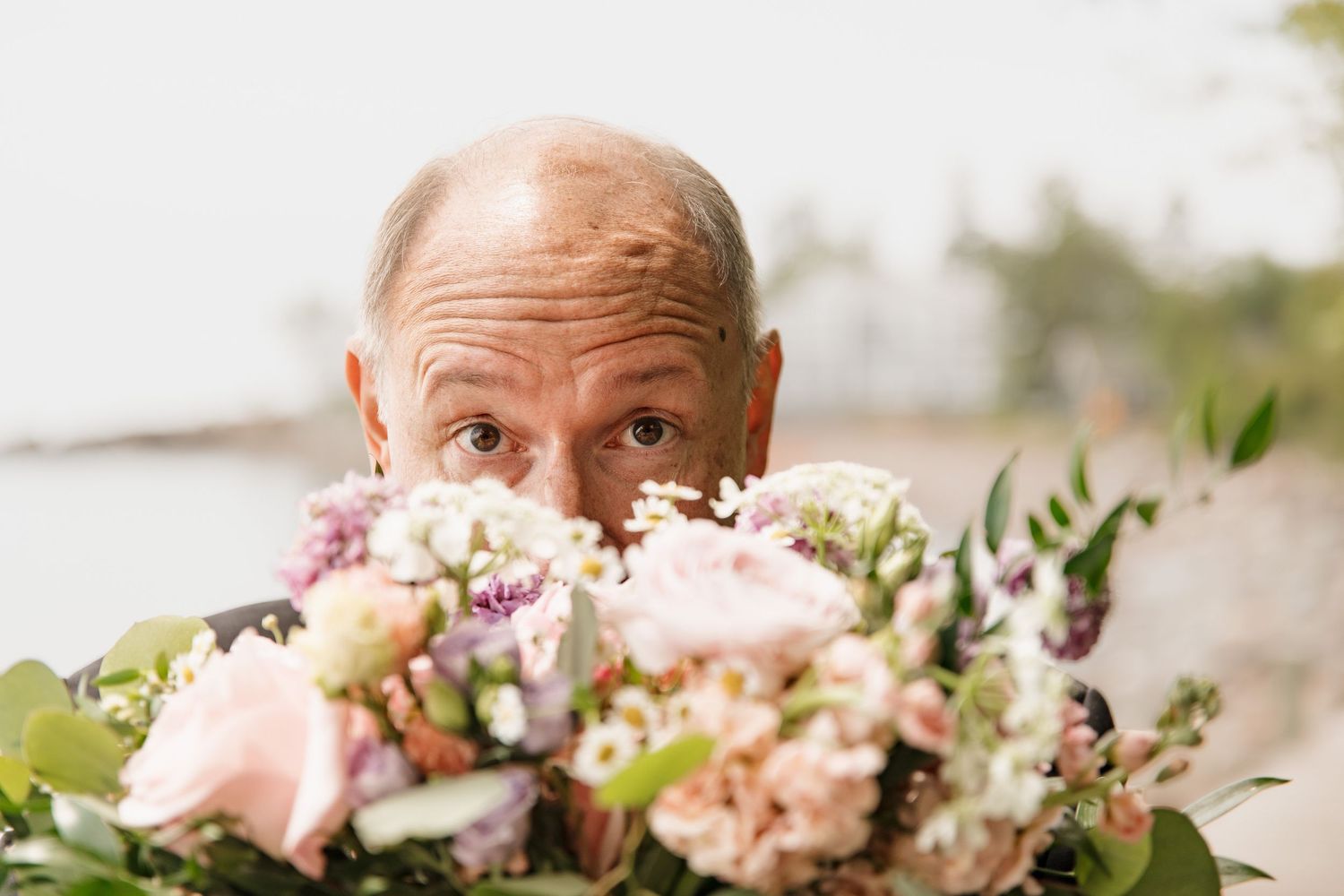 Bald groom playfully peeking over a lush pink and white wedding bouquet outdoors near the water.