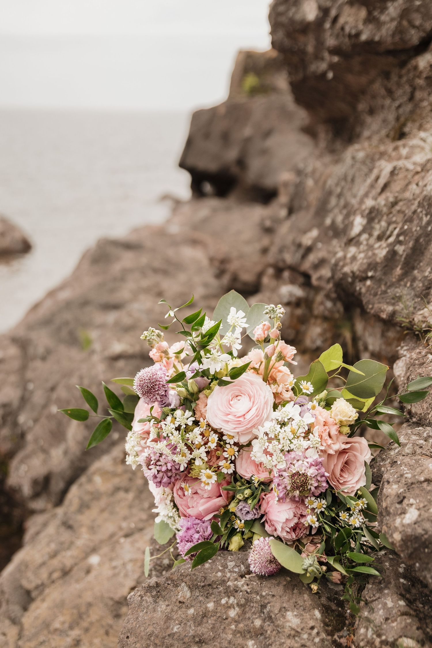 A romantic bridal bouquet of pink roses and wildflowers resting on rocky coastal cliffs with ocean in background.