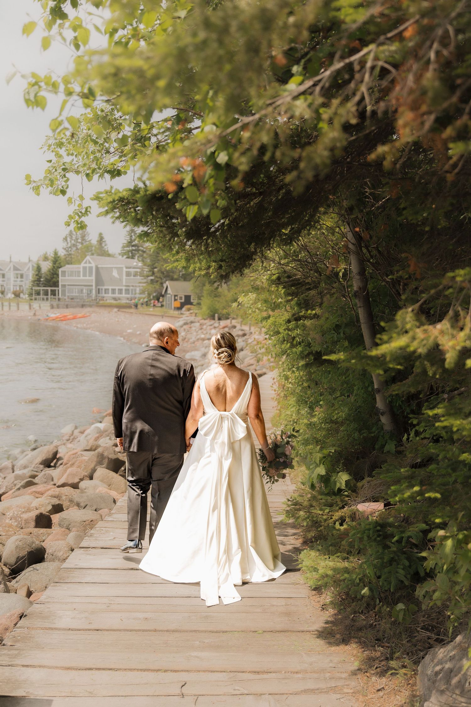 Bride and groom walking hand in hand along a wooden boardwalk beside a rocky tree-lined waterfront.