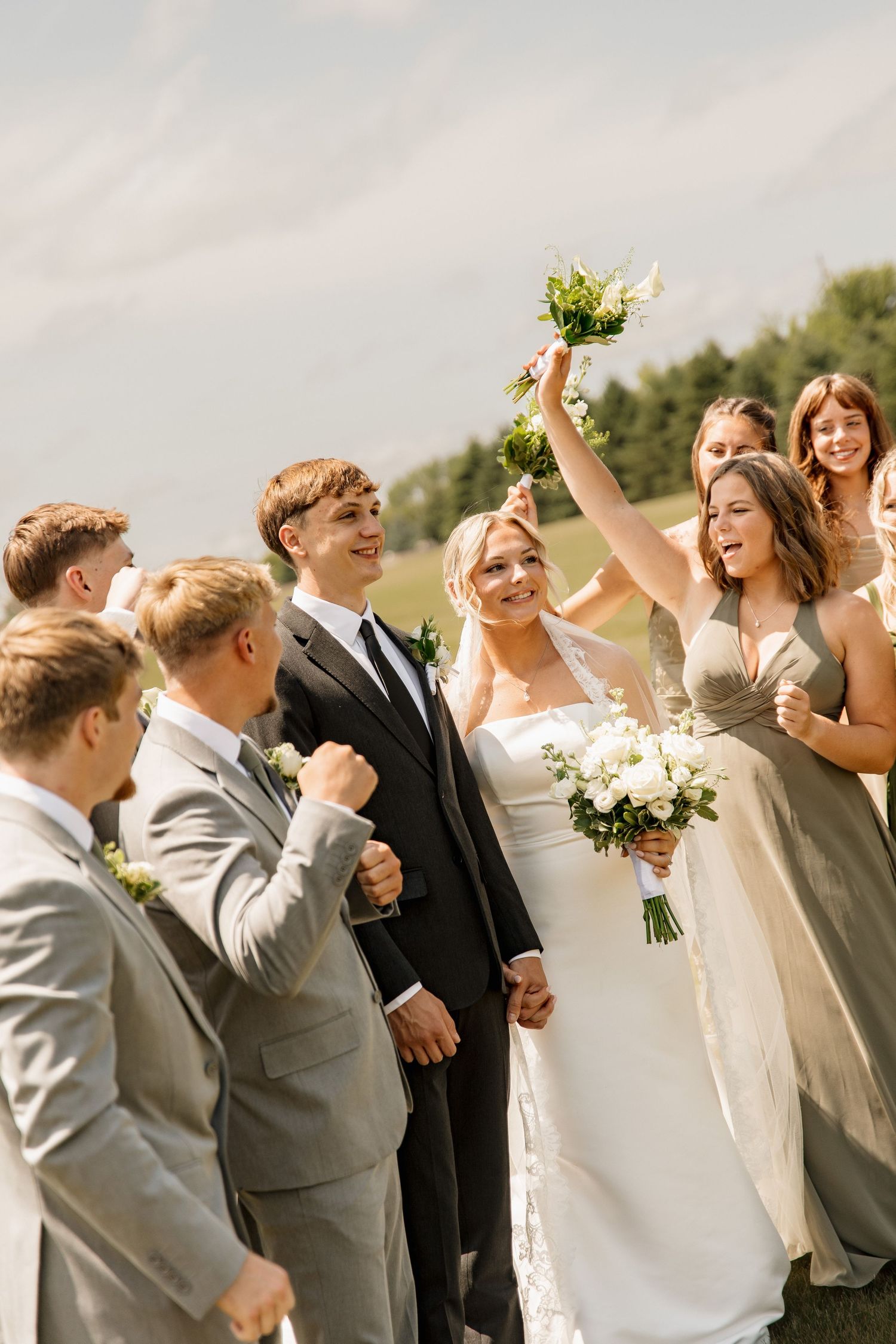 Bride and groom celebrating with wedding party in sage green dresses and gray suits outdoors.