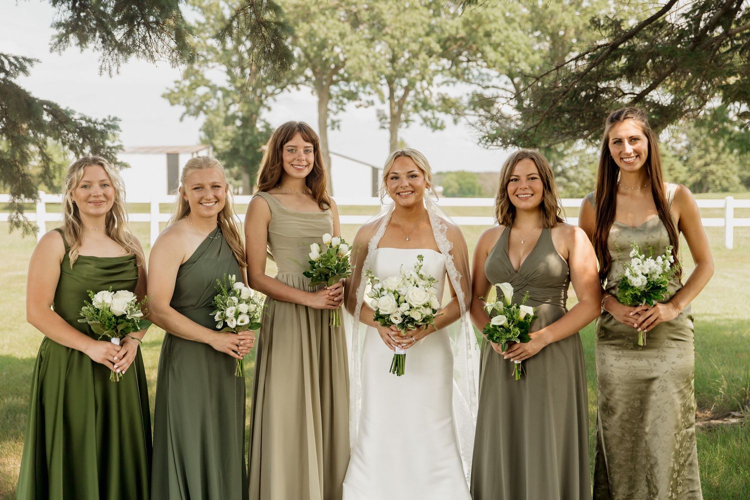 Bride in white gown with five bridesmaids in sage green dresses holding white bouquets outdoors.