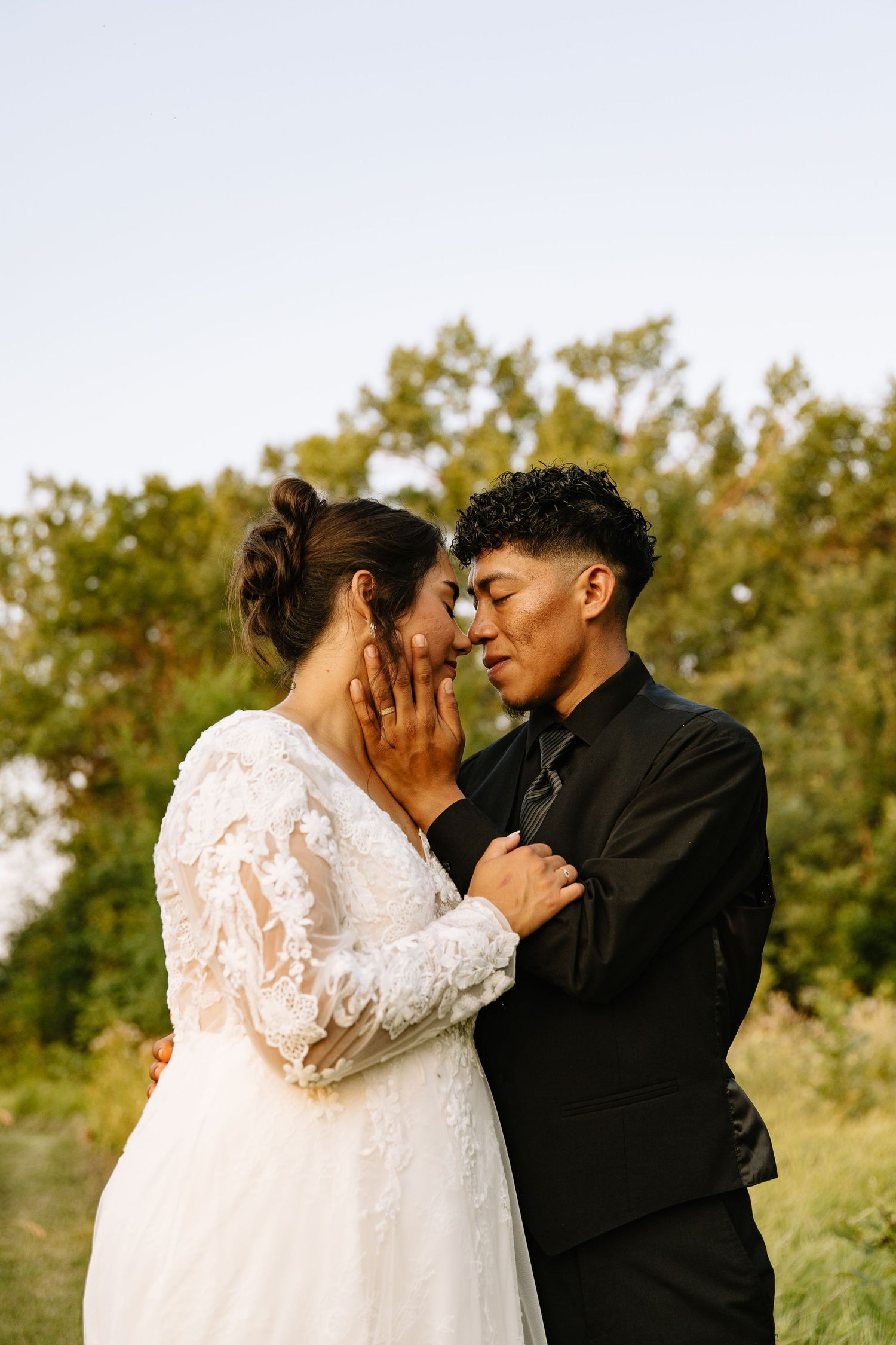 Bride in lace gown and groom in black suit share an intimate moment outdoors with trees in background.
