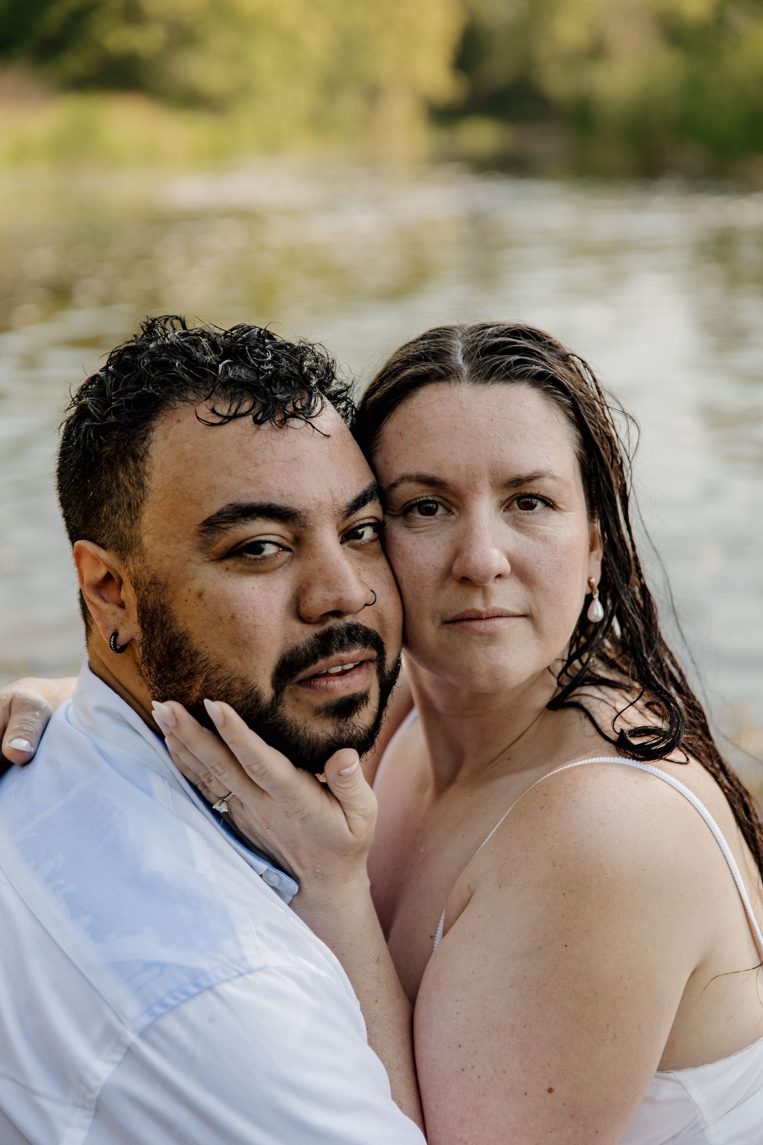 A couple poses intimately near a river, the woman resting her cheek against the bearded mans face outdoors.