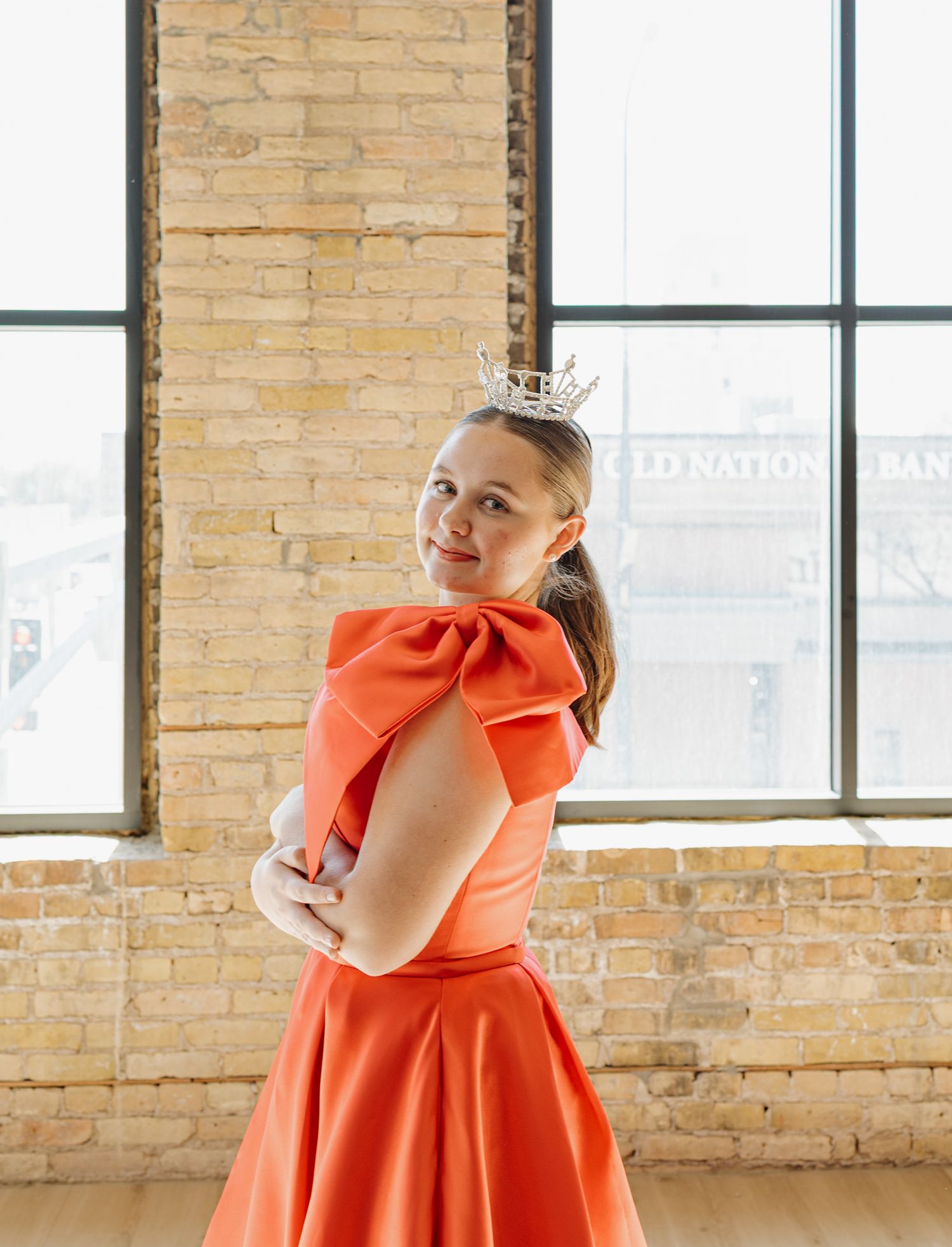 Young woman in orange bow dress with crown posing in sunlit brick-walled studio space.