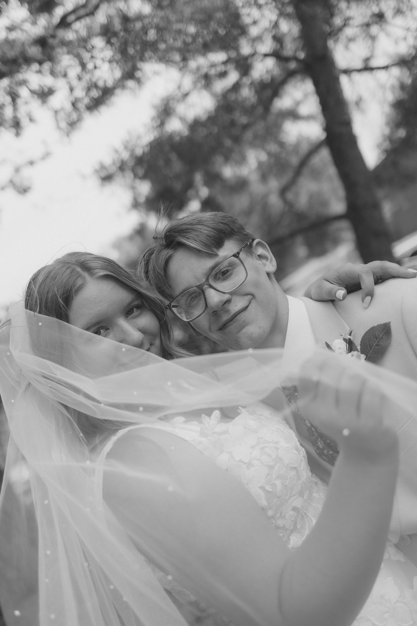 Black and white photo of a bride and groom posing together under a flowing wedding veil outdoors.